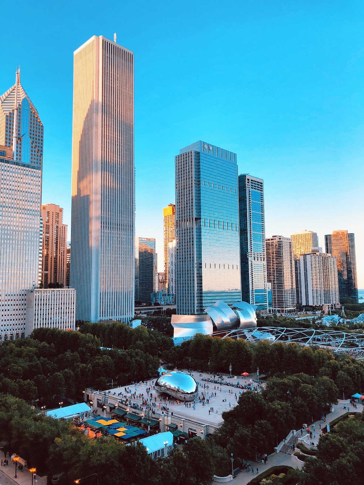 An aerial view of a downtown urban skyline featuring multiple modern high-rise buildings and skyscrapers against a clear blue sky. In the foreground, a public park area contains a large reflective bean-shaped sculpture and a contemporary curved metallic pavilion structure, surrounded by green trees and pathways with visitors visible as small figures in the plaza. The scene is photographed during golden hour lighting, casting warm tones on the building facades.
