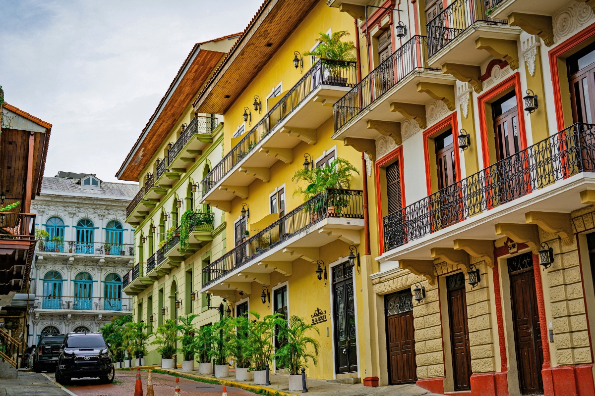 A row of multi-story colonial-style buildings features bright yellow and cream-colored facades with decorative balconies, ornate wrought iron railings, and red-trimmed windows and doors. The architecture includes terracotta roof tiles, carved corbels, and wall-mounted lanterns, with potted palm plants in white containers lining the street level. Additional historic buildings with turquoise accents are visible in the background, and vehicles are parked along the cobblestone or paved street.