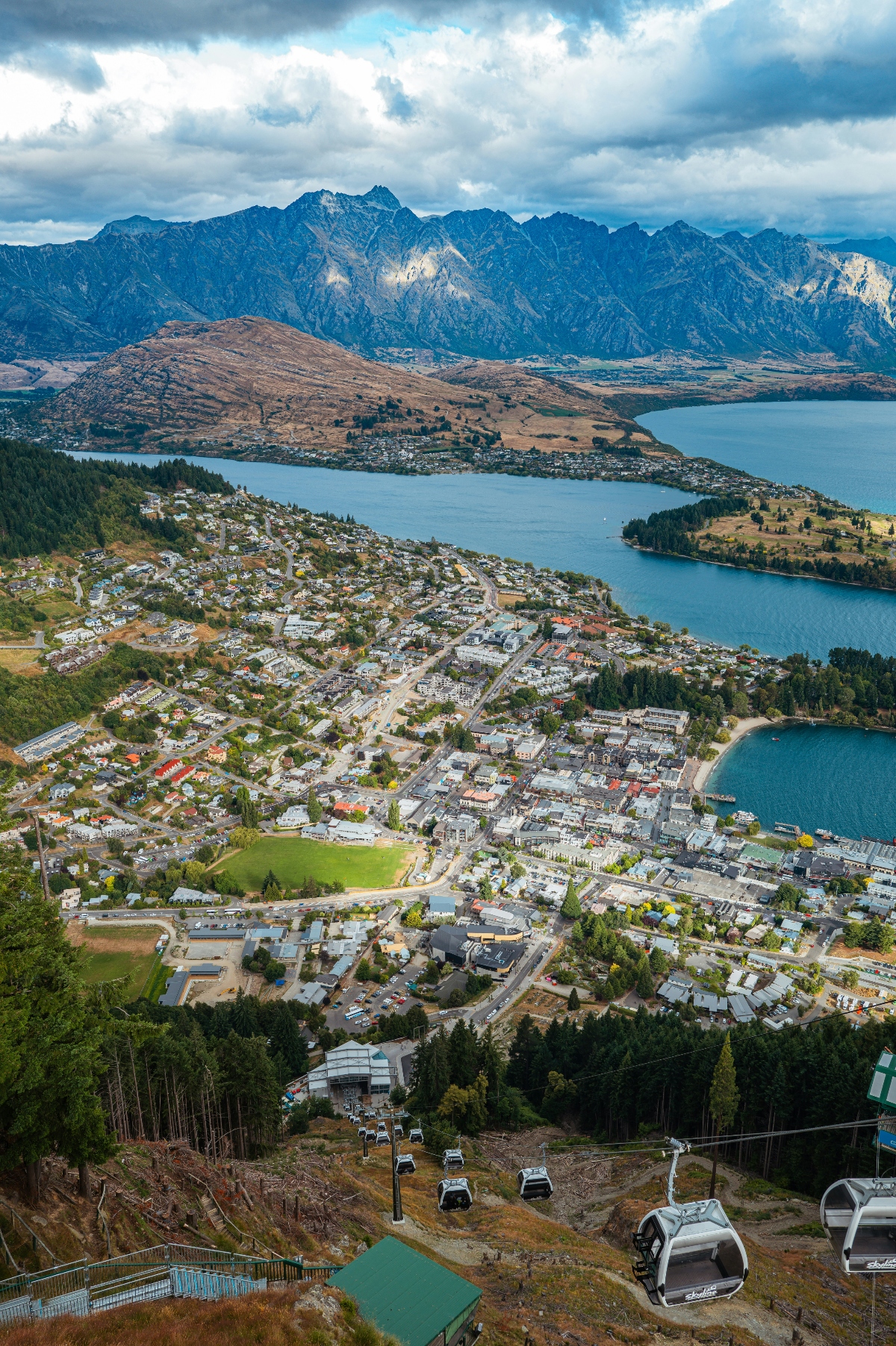 An aerial view shows a lakeside town nestled between a large blue lake and a dramatic jagged mountain range in the background, with dramatic clouds overhead. In the foreground, a gondola cable car system descends a forested hillside toward the densely built town center along the waterfront. The landscape features a mix of urban development, green spaces, forested areas, and brown hills across the lake, creating a scenic alpine setting.