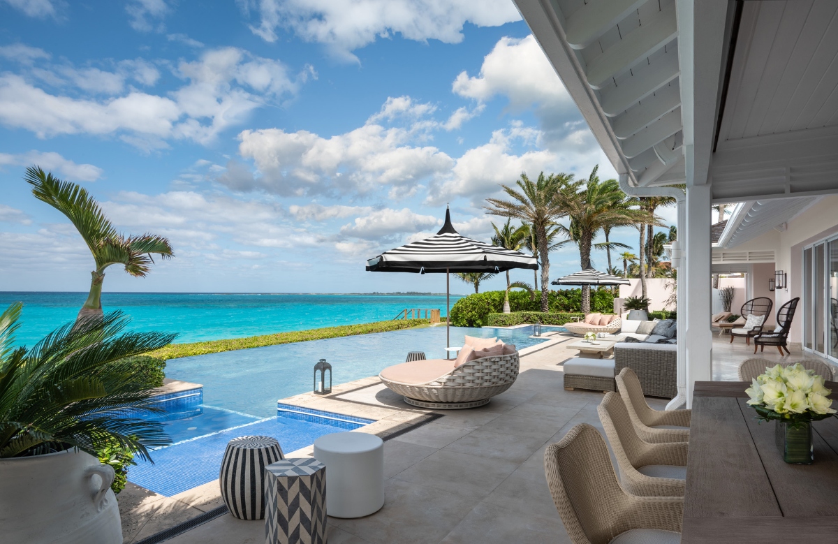A luxurious oceanfront terrace at Ocean Club in the Bahamas, ideal for a family vacation with adult children, features an infinity pool with pristine blue water that seamlessly blends into the stunning turquoise Caribbean Sea beyond. The elegant outdoor living space includes a white-covered pavilion with wicker furniture, striped umbrellas, and tropical palm trees set against a backdrop of brilliant blue skies and fluffy white clouds.