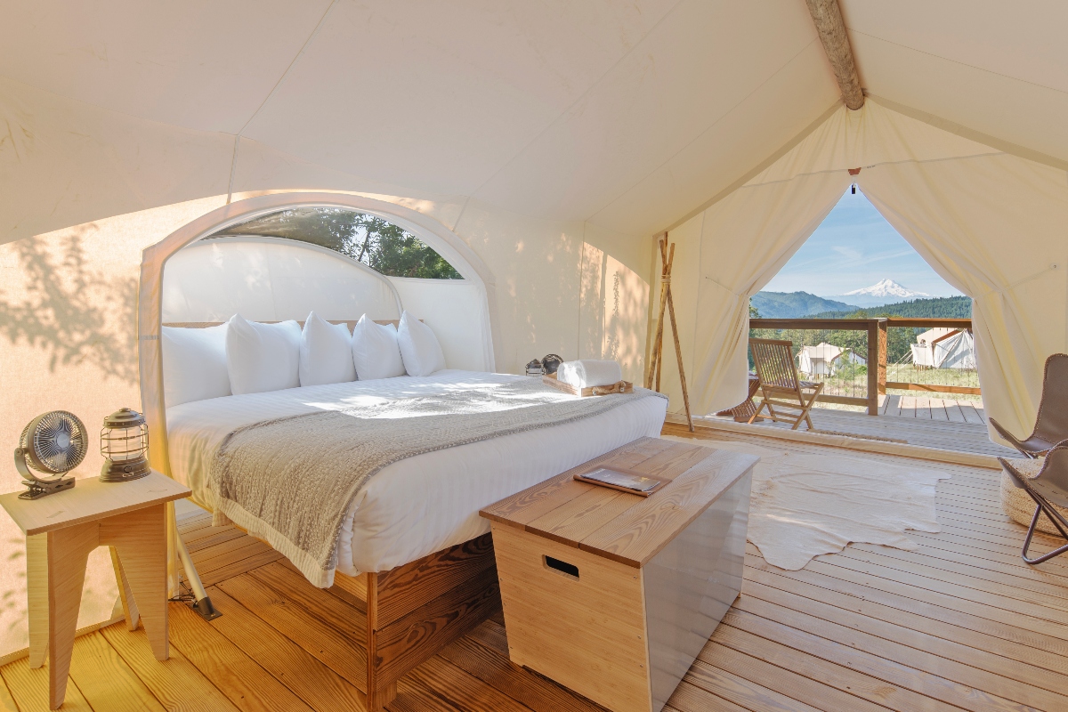 A luxury safari-style tent interior at Under Canvas Columbia River Gorge features a comfortable bed with crisp white linens and pillows, surrounded by warm wood flooring and furnishings including a wooden bench and side table. The spacious canvas accommodation opens to reveal stunning mountain views and outdoor seating on a private deck, creating an upscale glamping experience for a family vacation with adult children.