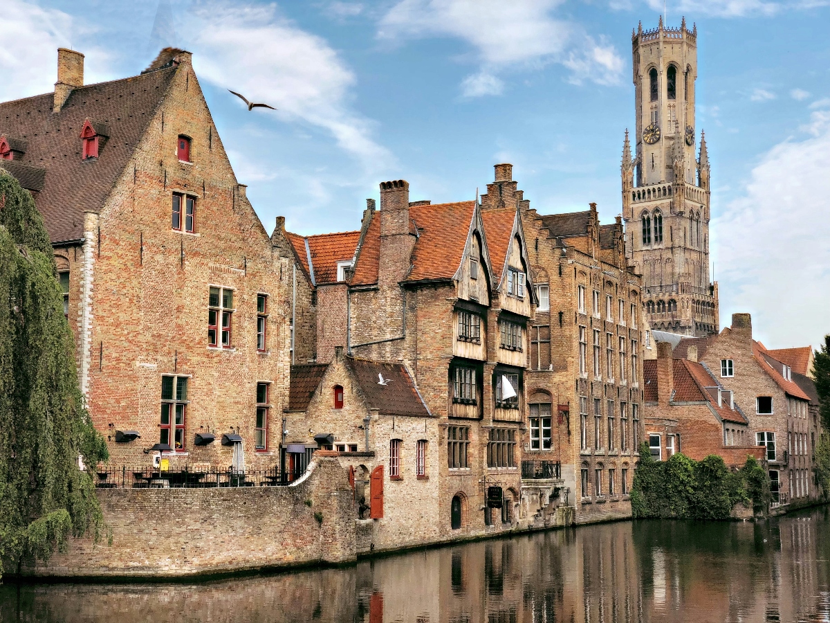 A row of historic brick buildings with stepped gables and orange-tiled roofs lines a calm canal, their facades reflected in the water below. A tall Gothic bell tower with ornate stonework rises prominently above the roofline on the right side of the scene. A bird flies through the blue sky dotted with white clouds above the medieval architecture.