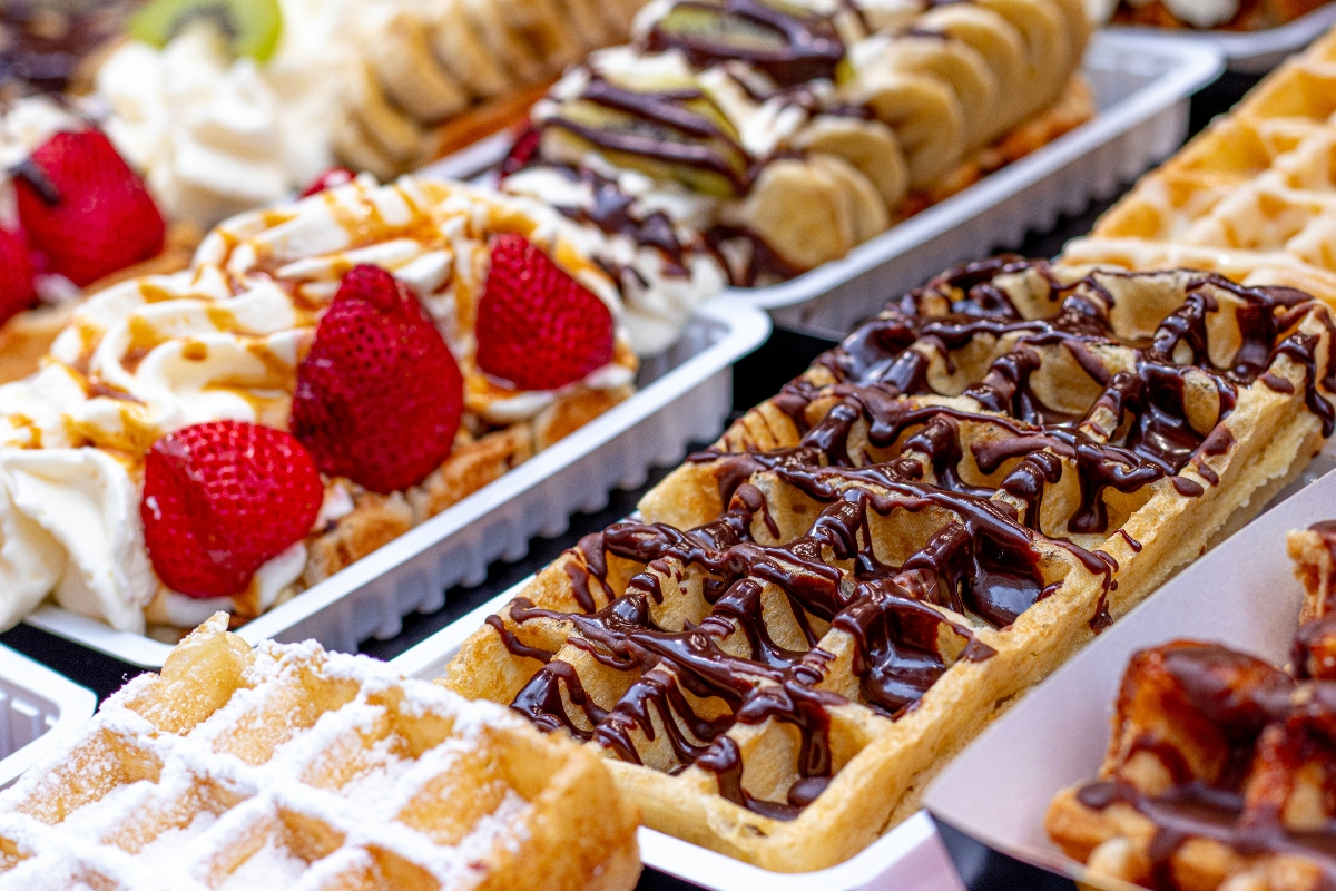 A display of golden Belgian waffles arranged in metal trays, each topped with different combinations of toppings including whipped cream, fresh strawberries, sliced bananas, chocolate drizzle, caramel sauce, and powdered sugar. The waffles feature the characteristic deep grid pattern and are presented in an overlapping arrangement. The toppings are artfully applied in decorative patterns across the various waffles.