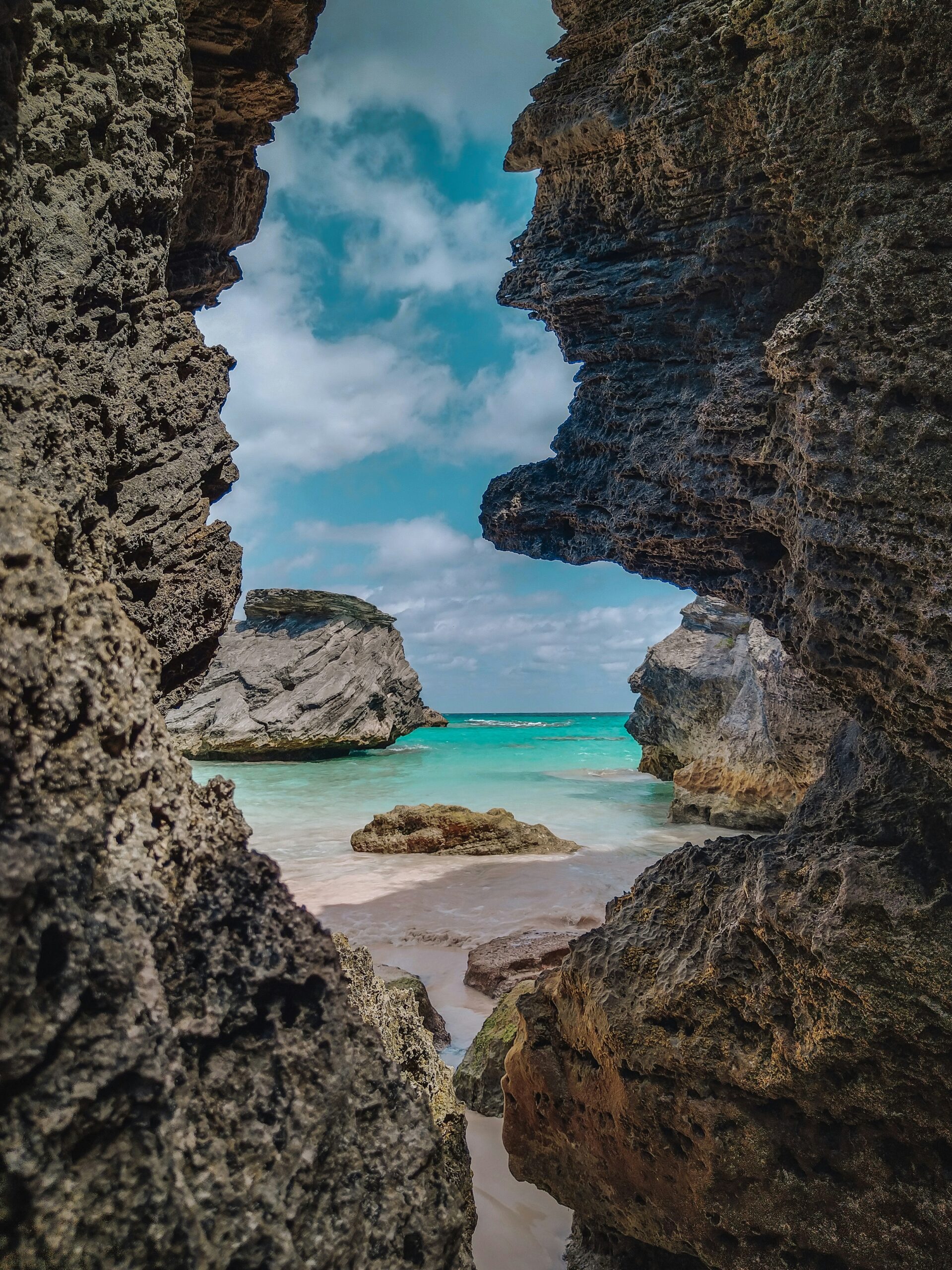 A pink sand beach on Bermuda is seen through a gap between two rough stacks of dark rock. Beyond is the turquoise sea and the blue sky.