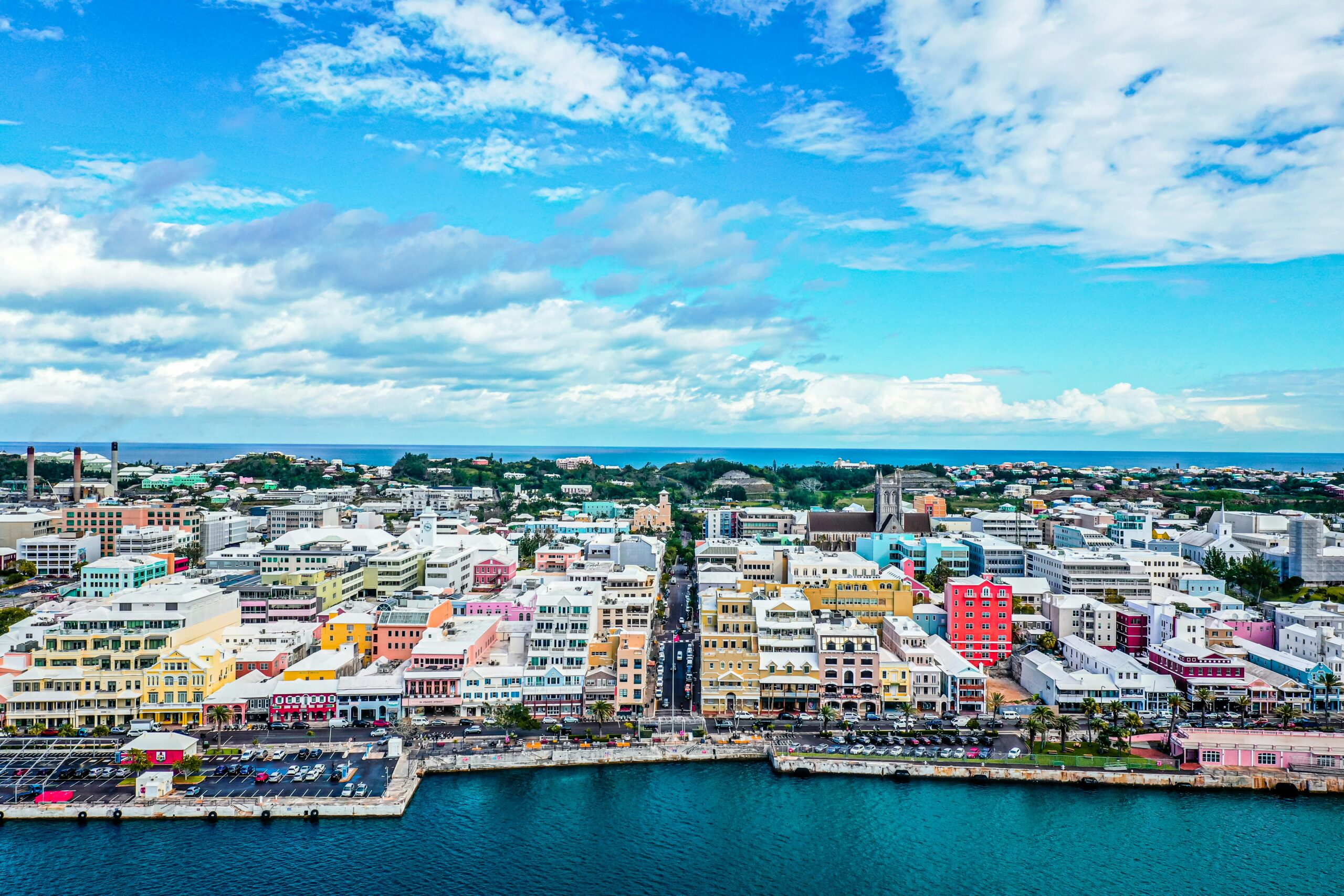 An aerial view of the waterfront city of Hamilton, Bermuda, showing a harbor with dark blue water in the foreground and a dense collection of colorful buildings extending inland. The buildings are predominantly multi-story structures in various colors including pink, yellow, blue, white, orange, and green, creating a vibrant urban landscape.