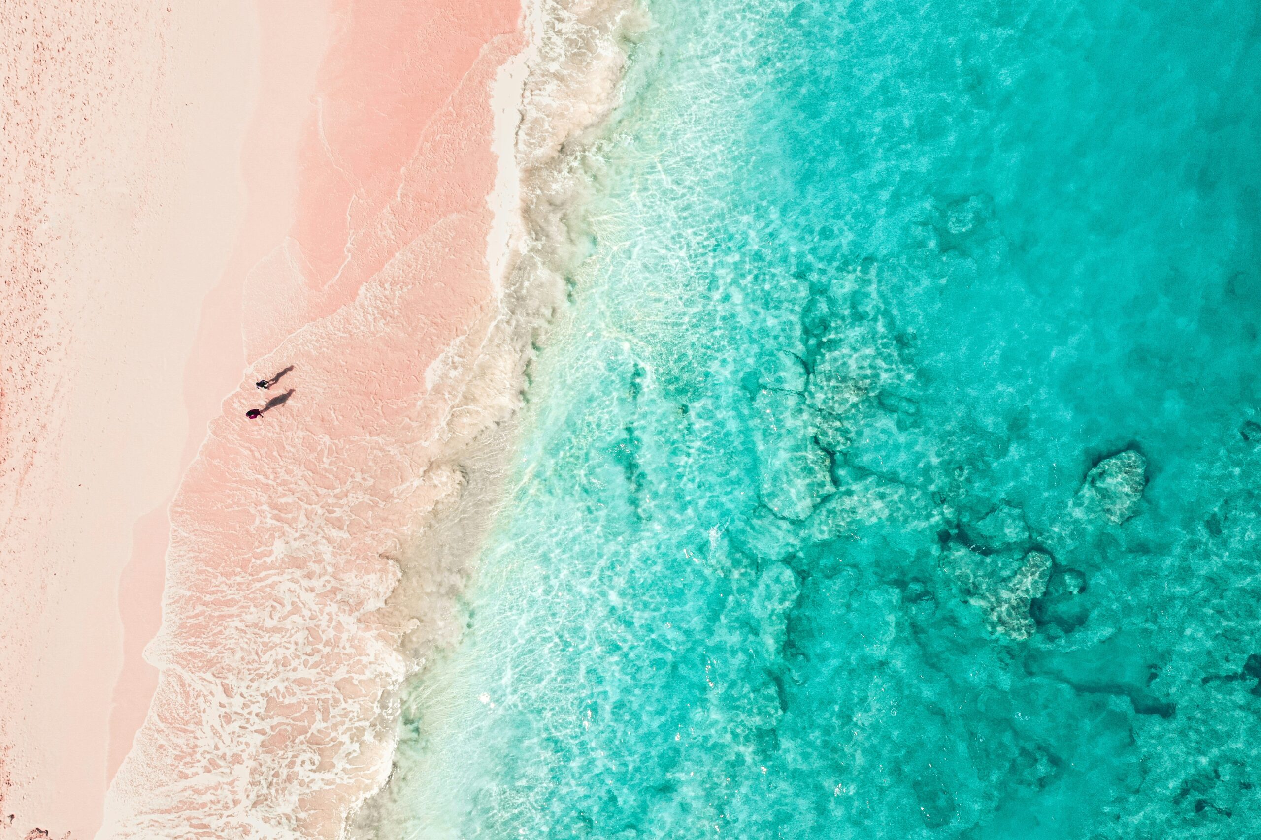 An aerial shot shows two people walking far below on a pink sand beach in Bermuda. The beach occupies the left side of the image; the right side shows the turquoise ocean shallows with rocks under the waves.