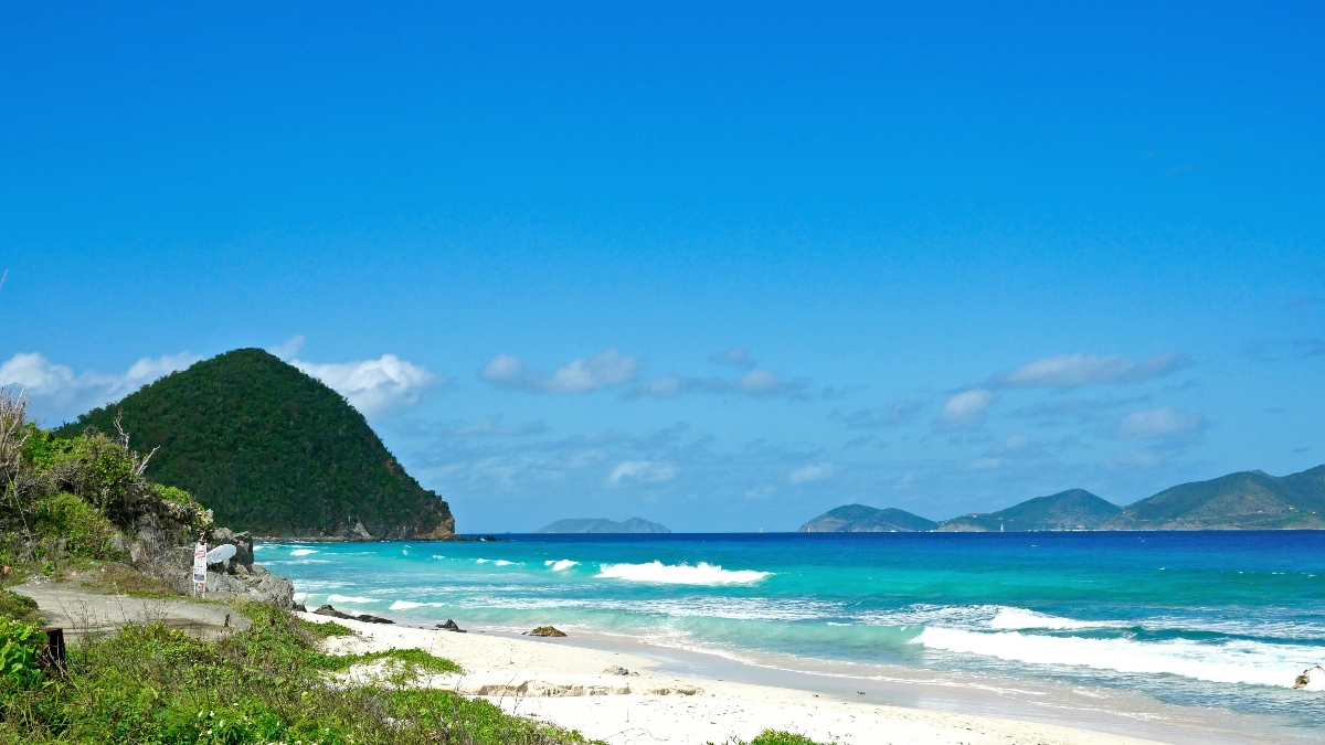 A white sandy beach with turquoise ocean waters and rolling waves, bordered by green vegetation on the left and a large, densely forested rocky hill jutting into the sea. Additional mountain peaks are visible across the water in the distance under a bright blue sky with scattered white clouds.
