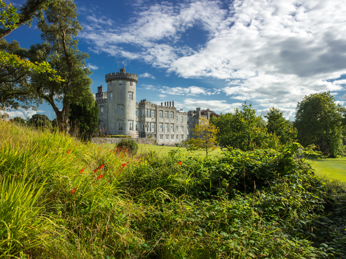 A stone castle with a prominent round tower and crenellated walls sits amid landscaped grounds, with a multi-story manor building extending from the main structure. The foreground features overgrown vegetation including tall grasses and red flowering plants, while mature trees frame the scene on the left side. The sky shows a mix of white clouds and blue areas above the building and surrounding green lawns.