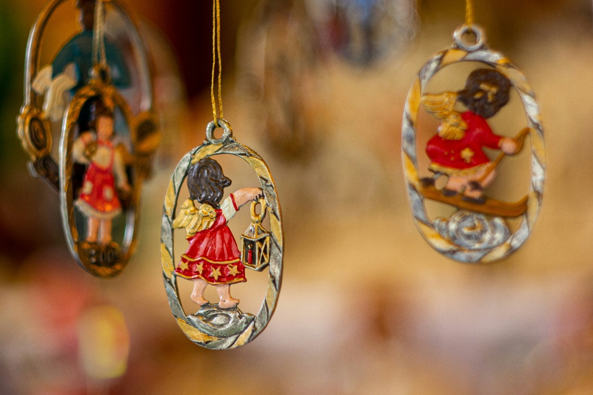Three hand-painted Christmas ornaments hang from gold cords against a blurred warm-toned background. The ornaments are oval-shaped with decorative twisted edges and feature miniature scenes with figures in traditional clothing alongside animals, including a central ornament depicting a person in a red dress holding a lantern with a bear figure. The detailed craftsmanship and style suggest traditional European Christmas market decorations.