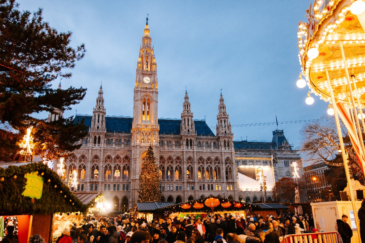 A Gothic Revival building with a prominent central spire and clock tower stands illuminated against the evening sky, with four smaller matching spires flanking the main structure. In front of the building, a large Christmas market bustles with crowds of people among wooden market stalls, featuring a tall decorated Christmas tree and carnival rides including a brightly lit carousel on the right side.