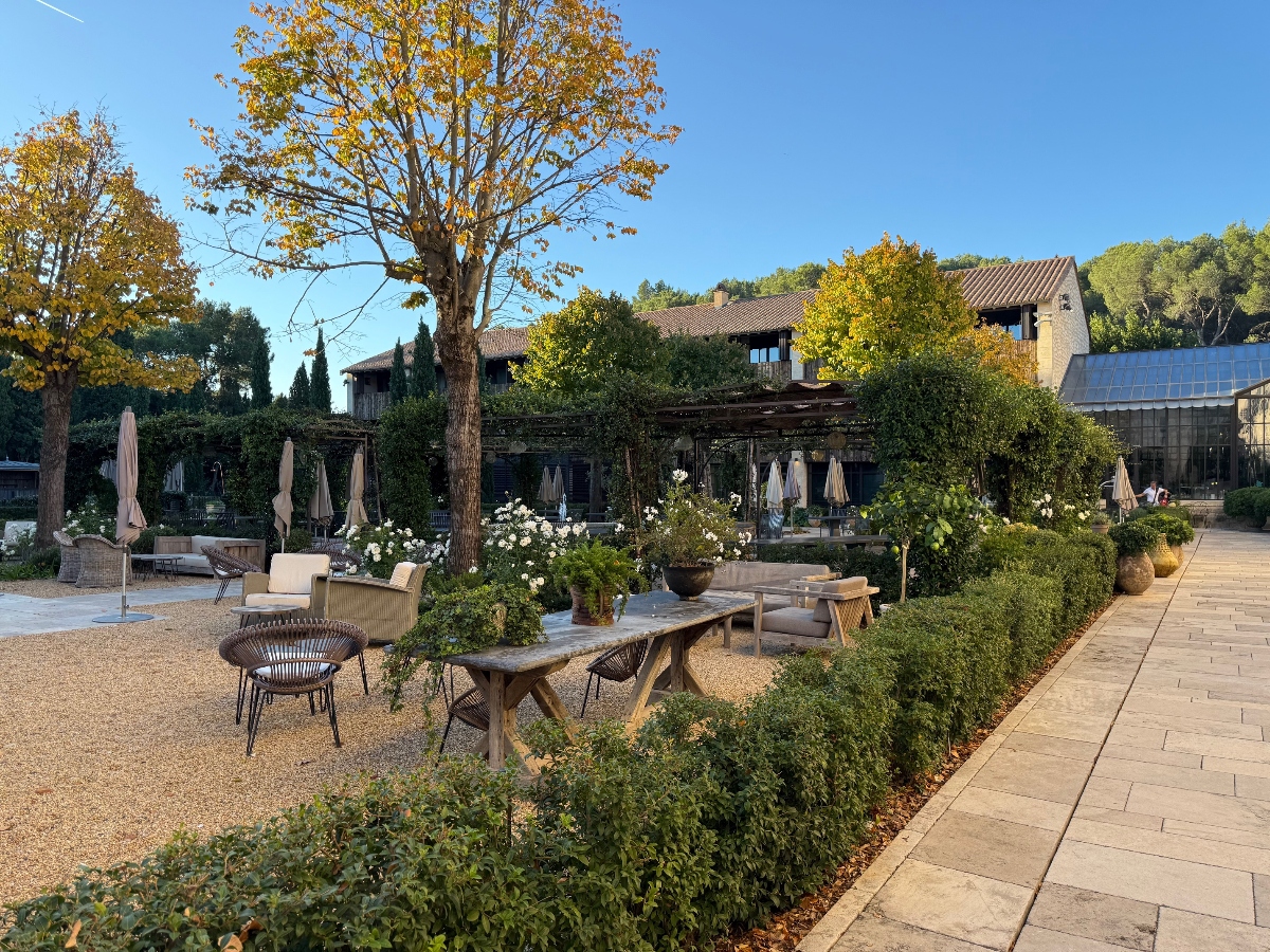 A resort courtyard features rustic wooden tables and wicker chairs arranged on gravel pathways, with a large tree displaying autumn foliage at the center. Stone buildings with terracotta roofs surround the space, connected by a covered pergola structure, while manicured hedges and potted plants line the walkways. Furled umbrellas and lounge furniture are visible in the background, and a stone paved path runs along the right side of the courtyard.