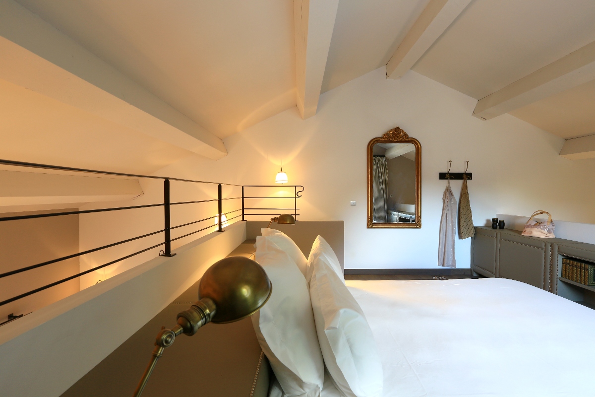 A loft bedroom features a bed with white linens and a brass reading lamp in the foreground, with vaulted ceilings supported by exposed wooden beams. A black metal railing runs along the edge of the mezzanine level, overlooking a lower floor space. The far wall displays a gold-framed mirror, wall hooks with hanging robes, and a gray dresser beneath a sloped ceiling.