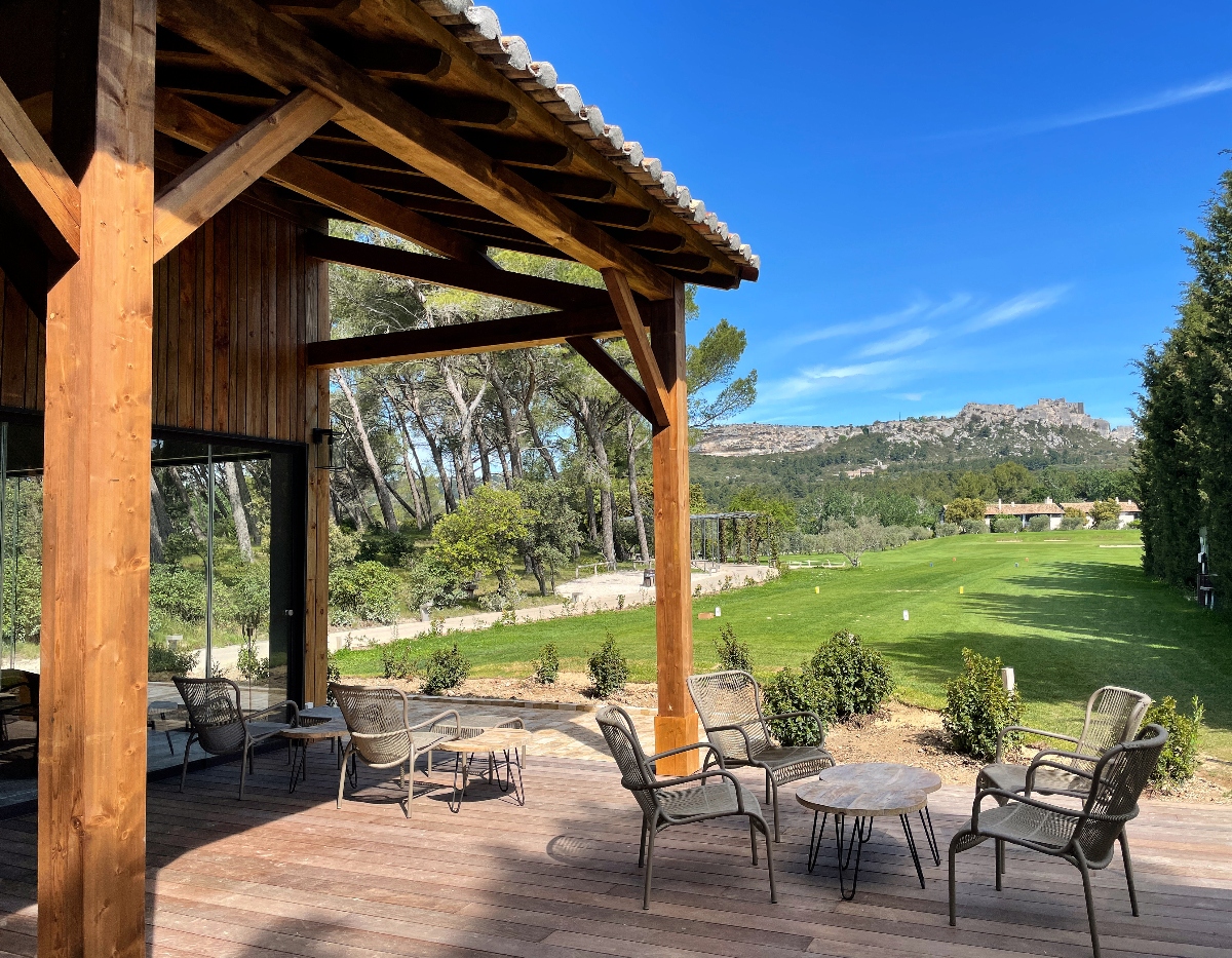 A covered wooden terrace with exposed timber beams and a tile roof overlooks a golf course, with several dark wicker chairs and round tables arranged on a wooden deck. Beyond the manicured green fairway, mature trees and residential buildings are visible in the middle distance. A rocky mountain ridge rises against a blue sky in the background.