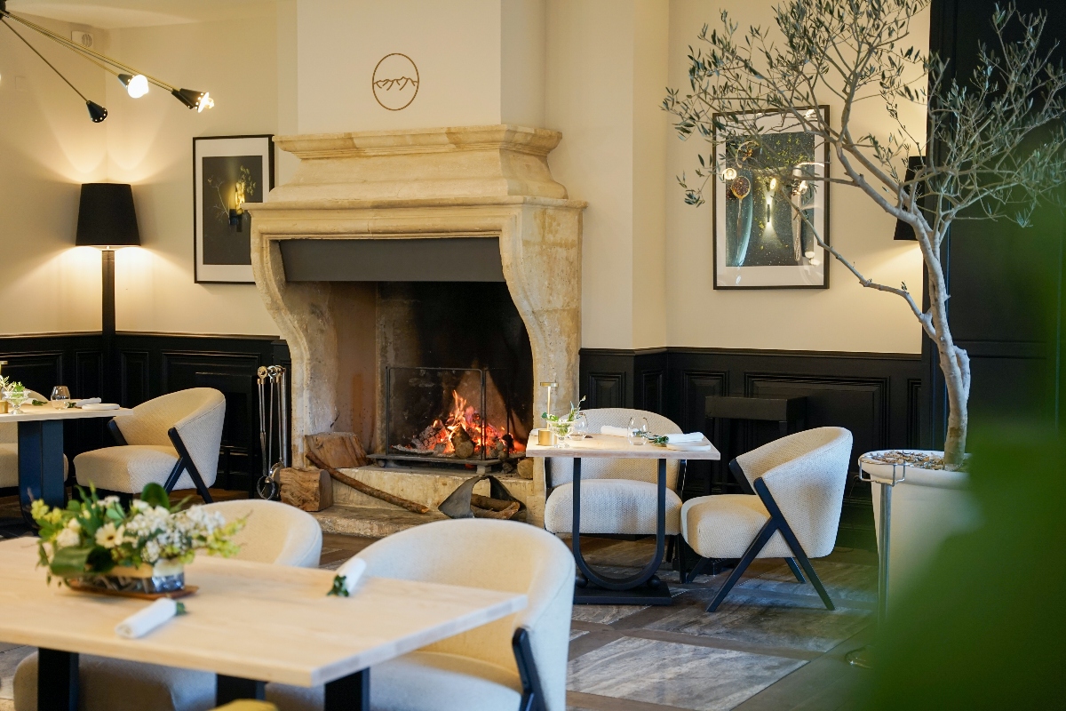 A restaurant dining room features a large stone fireplace with an active fire as its centerpiece, surrounded by white-clothed tables with cream upholstered chairs that have black angular frames. The space has dark wood wainscoting on the lower portion of cream-colored walls, with framed artwork, lighting fixtures, and a decorative olive tree branch positioned near the right side of the room. Firewood is stacked beneath the carved stone mantelpiece.