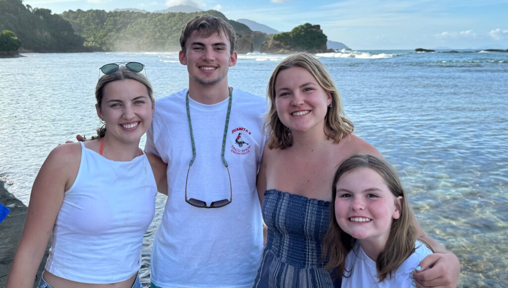 Four young people smile together for a group photo on a scenic coastline, with the young man in the center wearing a white t-shirt and sunglasses around his neck, flanked by three young women in summer clothing. Behind them stretches a beautiful coastal landscape featuring calm blue-green waters, forested hills and rocky outcroppings, with mountains visible in the distance under a partly cloudy sky.