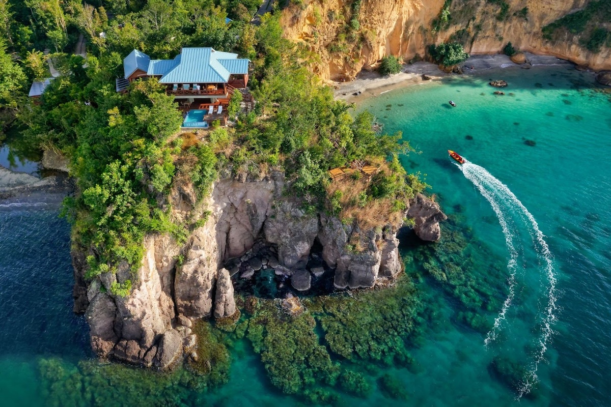 An aerial view shows a luxurious villa with distinctive blue roofing perched on dramatic rocky cliffs overlooking crystal-clear turquoise waters, surrounded by dense tropical vegetation. A boat cuts through the vibrant emerald and blue waters near a small secluded beach, with coral formations visible beneath the surface and rugged limestone cliffs creating a stunning coastal landscape.