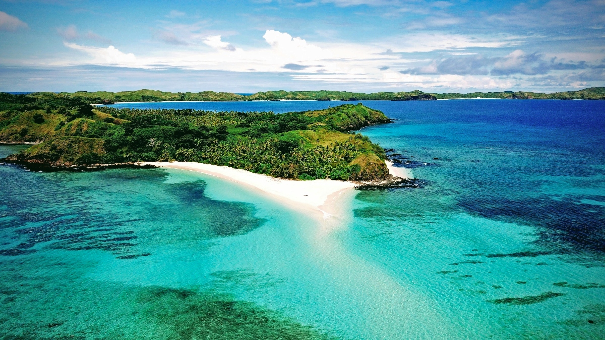An aerial view shows a tropical coastline with a distinctive white sand spit or sandbar extending into crystal-clear turquoise and blue waters of varying depths. The landscape features lush green hills covered in dense vegetation, curved beaches, and a partly cloudy sky over the ocean horizon.