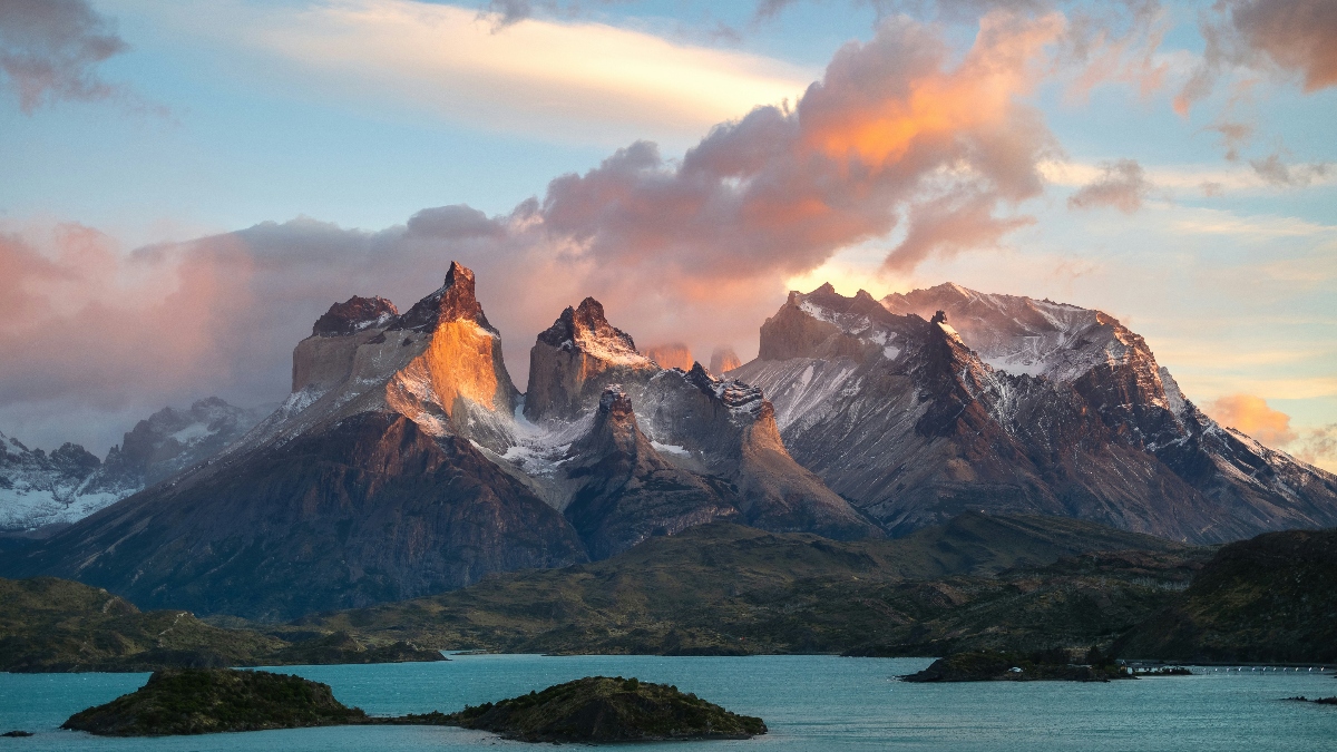 A dramatic mountain range with jagged, snow-capped granite peaks rises above a large turquoise lake, with several small islands visible in the foreground water. The scene is captured during golden hour lighting, with warm orange and pink hues illuminating the rocky spires against a cloudy sky.