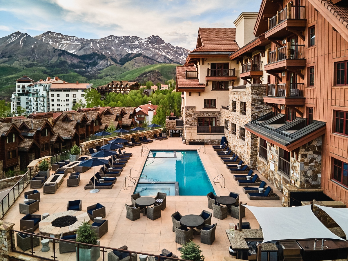 A luxury mountain resort features a large outdoor swimming pool and hot tub surrounded by lounge chairs and outdoor dining furniture on a stone patio. The resort buildings showcase a mix of architectural styles including stone, wood siding, and traditional alpine design elements, set against a backdrop of snow-capped mountains and green foothills. Multiple levels of accommodations and terraced outdoor spaces create an upscale mountain village atmosphere.