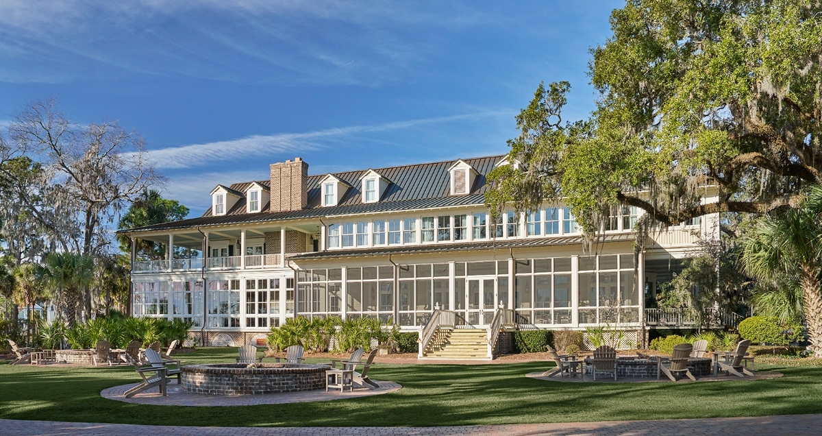 A large, multi-story brick building with colonial-style architecture features dormer windows, a prominent chimney, and an expansive screened porch spanning the ground level with wide stone steps leading up to it. The building is surrounded by manicured lawns and mature landscaping, including palm trees and a large oak draped with Spanish moss. In the foreground, there is a circular stone fire pit area on the grass lawn under a partly cloudy blue sky.