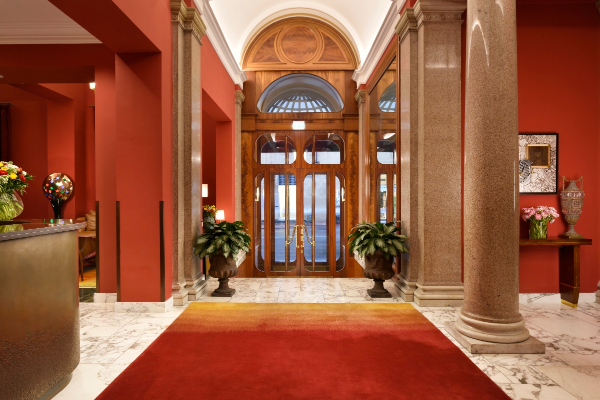 A grand hotel lobby features red walls, classical stone columns, and an ornate arched entrance with wooden doors topped by a decorative dome. A red carpet runner leads across white marble flooring to the entrance, which is flanked by large decorative planters and framed by elaborate architectural details.
