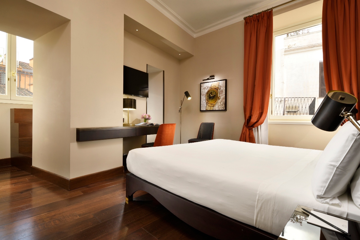 A modern hotel room features a bed with white linens, dark wooden floors, and a built-in desk area with an orange chair positioned near windows with orange curtains. The room has cream-colored walls, a wall-mounted television, and opens to a balcony overlooking neighboring buildings with traditional architecture.