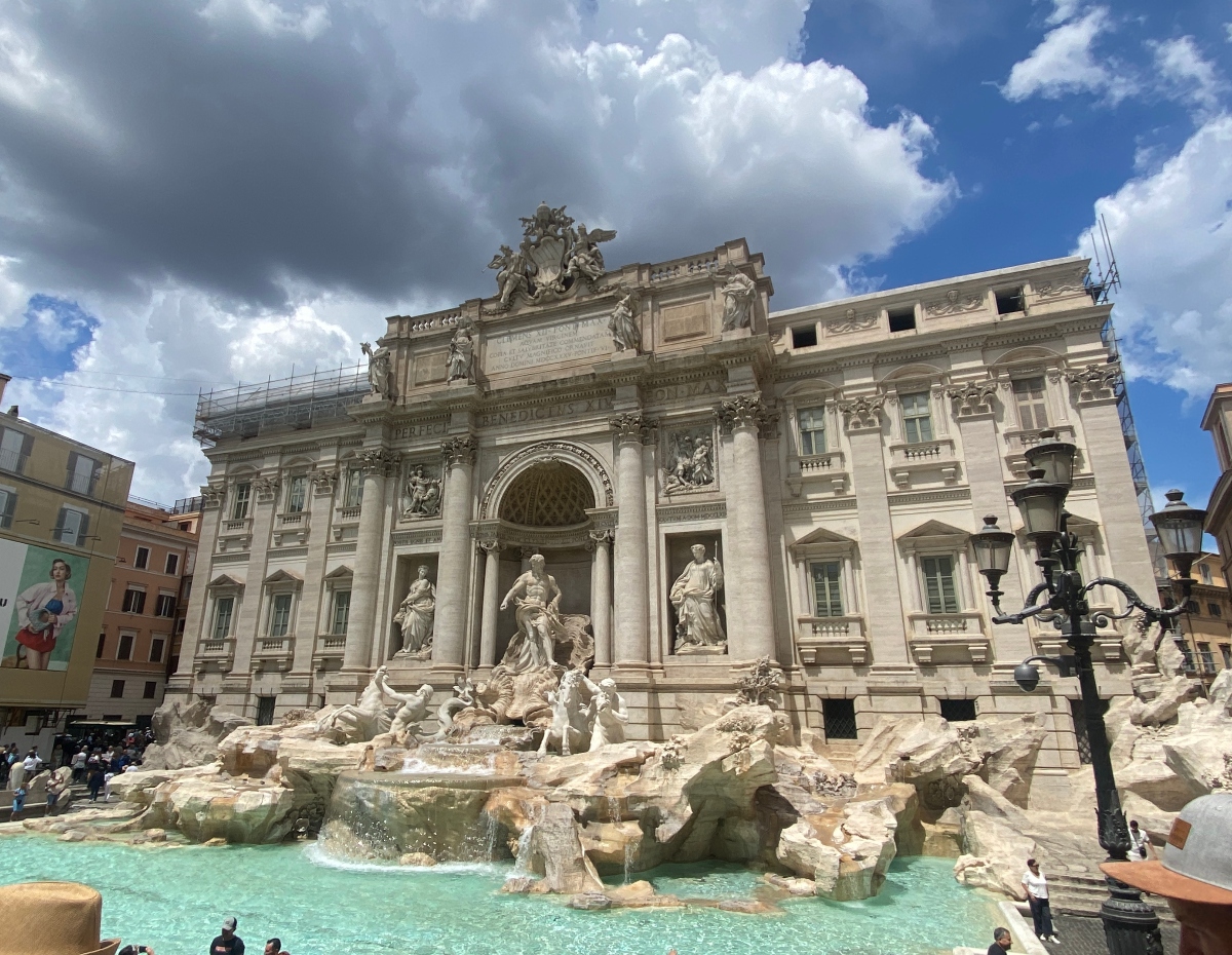 A large baroque fountain features elaborate white stone sculptures and architecture with a central arched niche containing carved figures, surrounded by classical columns and ornate decorative elements. Tourists gather around the fountain's turquoise water basin while dramatic clouds fill the blue sky above the surrounding historic buildings.