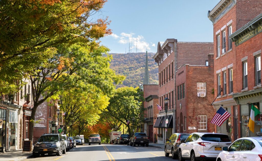 A tree-lined street runs through a historic downtown area with red brick buildings on both sides, cars parked along the curbs, and American flags displayed on several storefronts. In the background, a church steeple rises above the buildings with forested hills visible under a clear blue sky, while autumn foliage creates a canopy of green and golden leaves over the street.