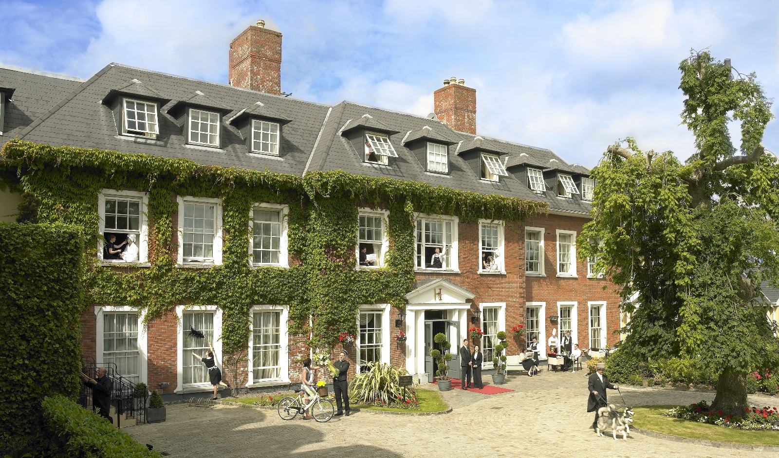 A large Georgian-style brick mansion with ivy-covered walls features multiple stories, dormer windows, and prominent brick chimneys under a partly cloudy sky. The building has white-framed windows throughout and a central entrance with white columns, while several people and dogs are visible on the curved gravel courtyard in front. Mature trees and landscaping surround the property, with a bicycle positioned near the entrance area.