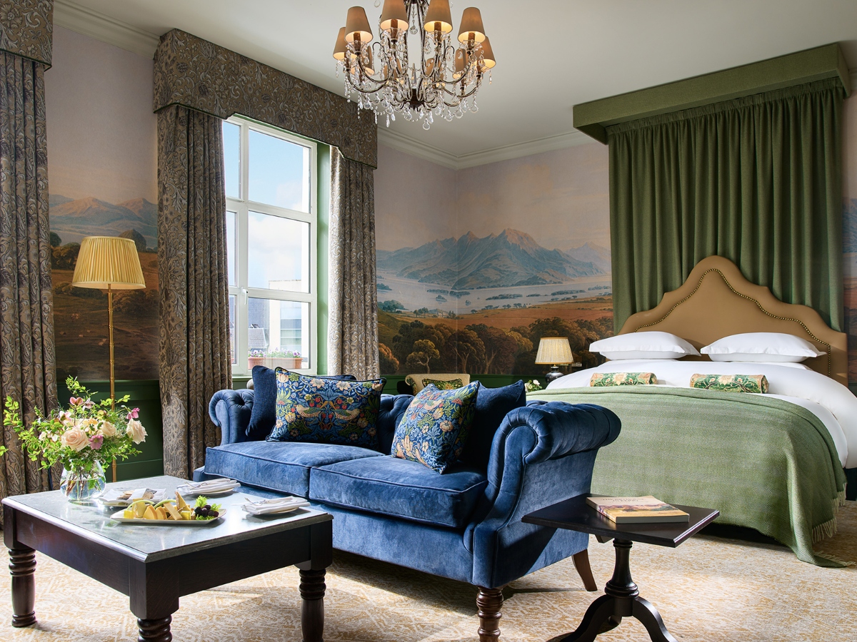 A luxurious hotel suite features a bed with white linens and beige upholstered headboard alongside a blue velvet sofa with patterned throw pillows positioned around a dark wood coffee table. The room is decorated with a large landscape mural depicting mountains and countryside scenes across the walls, green curtains, and patterned drapery framing a window. A crystal chandelier with fabric lampshades hangs from the ceiling, illuminating the traditionally appointed space with crown molding and refined furnishings.