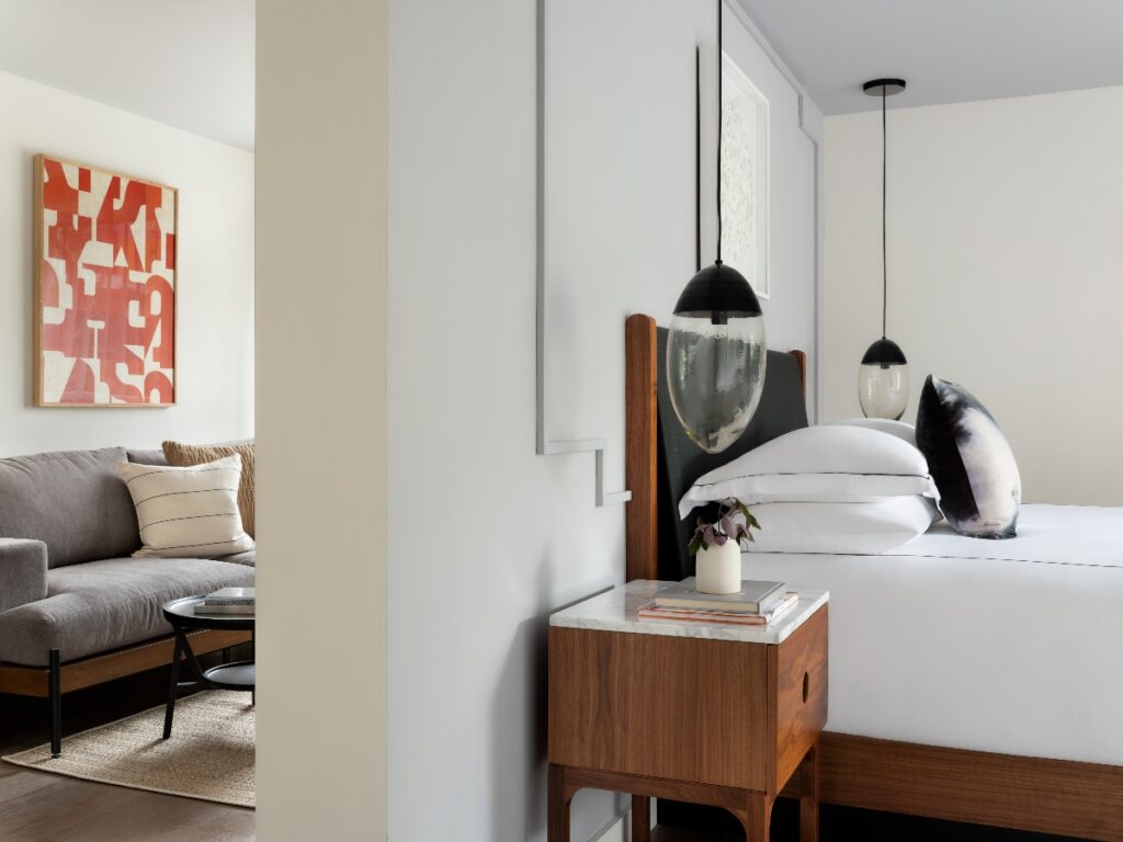 A modern bedroom and living room separated by a partial wall, featuring a white bed with crisp linens and black pendant lights beside a mid-century style wooden nightstand with a marble top. On the left, a gray sofa with striped pillows sits beneath abstract coral-toned wall art, while the bedroom wall displays a white-framed piece. The space has a minimalist aesthetic with neutral tones, wood accents, and clean lines throughout.