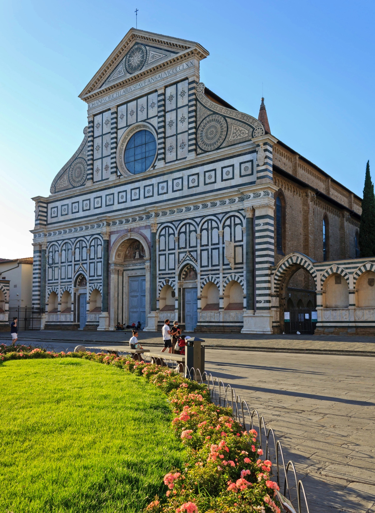 This image shows a large church or cathedral with a distinctive geometric facade featuring alternating horizontal stripes of white and dark green marble, topped by a triangular pediment with decorative circular motifs. The building displays Romanesque and Gothic architectural elements including arched doorways, a rose window, and ornate stonework, with several visitors visible on the stone plaza in front. In the foreground, there is a manicured lawn bordered by flower beds containing pink and coral-colored blooms, set against a clear blue sky.