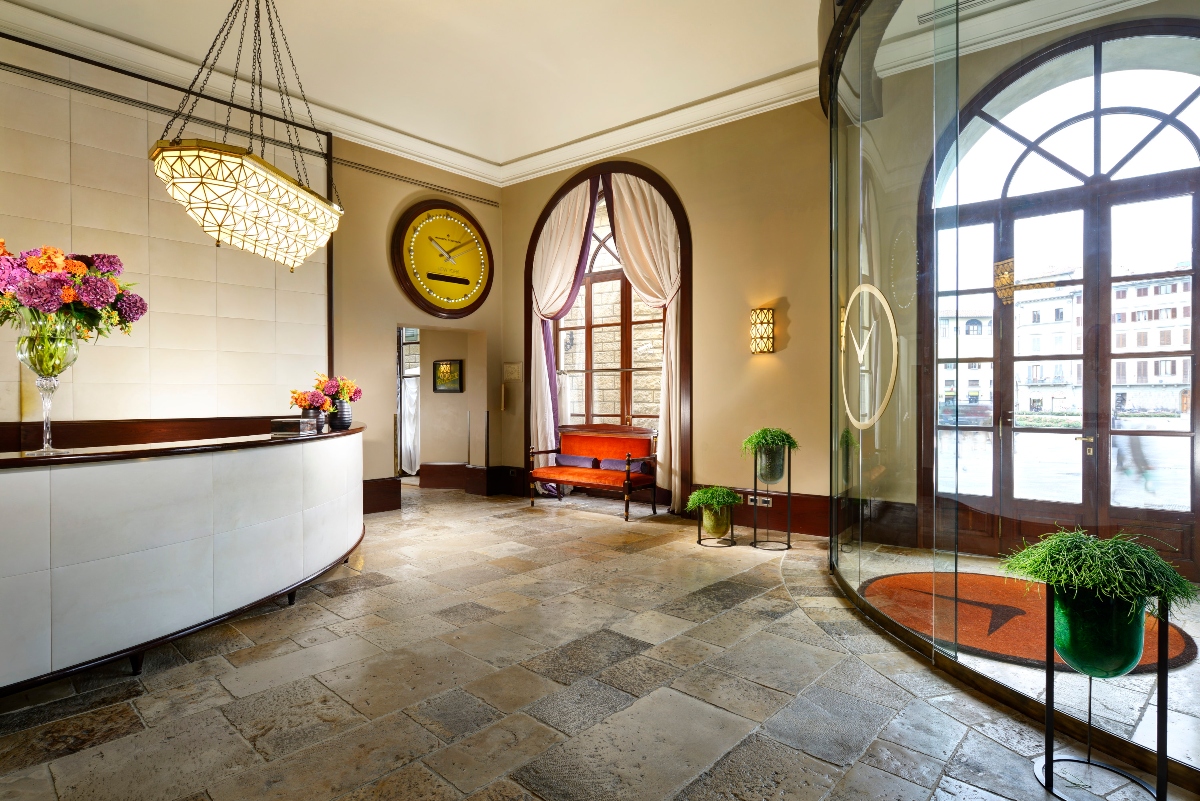 This image shows an elegant interior space with a curved white reception desk positioned beneath a large geometric crystal chandelier. The room features cream-colored walls with dark wood trim, arched windows with dark frames, and a mixed stone floor in various earth tones. A yellow wall clock, orange upholstered bench, potted plants, and a curved glass partition complete the sophisticated lobby or reception area design.