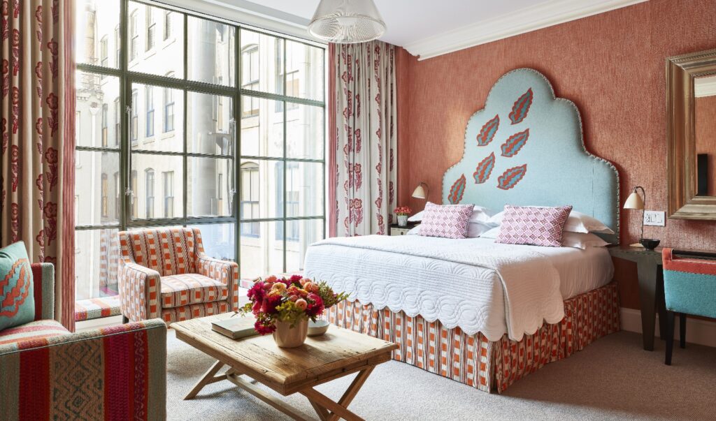 This hotel room features a distinctive curved blue headboard decorated with red paisley motifs, positioned against coral-colored walls with large multi-paned windows dressed in coordinating patterned curtains. The room includes a seating area with matching upholstered furniture in geometric patterns, a wooden bench at the foot of the bed displaying fresh flowers, and classical architectural details including crown molding and a decorative ceiling fixture.