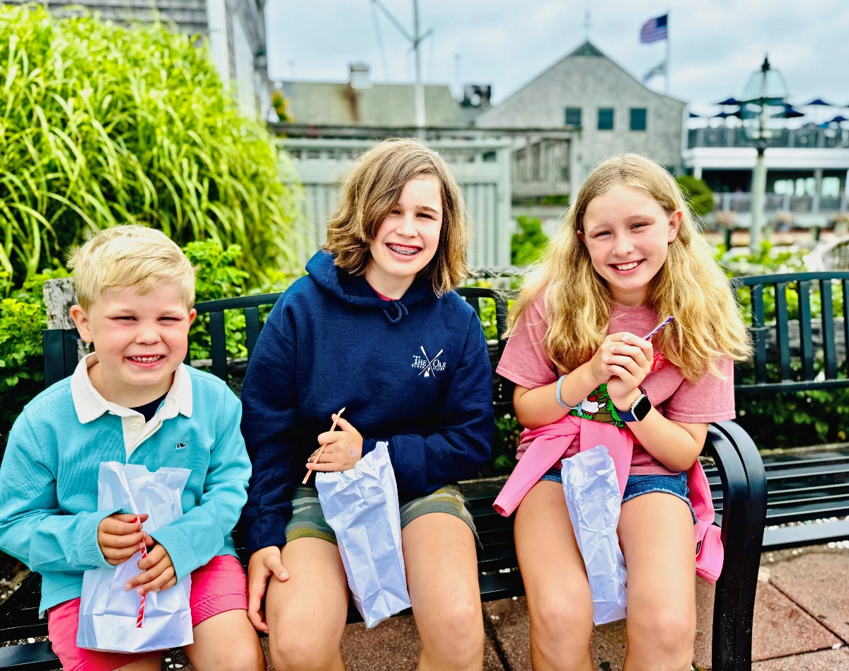 Three children sit together on a black bench, each holding white paper bags and smiling at the camera. The setting appears to be an outdoor waterfront area with lush green plants on the left side and buildings visible in the background.