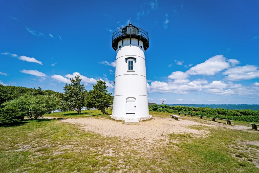 A white cylindrical lighthouse with a black lantern room and exterior railing stands on a grassy area surrounded by green trees and vegetation. The structure is set against a blue sky with white clouds, with the ocean visible in the background and wooden benches positioned nearby.