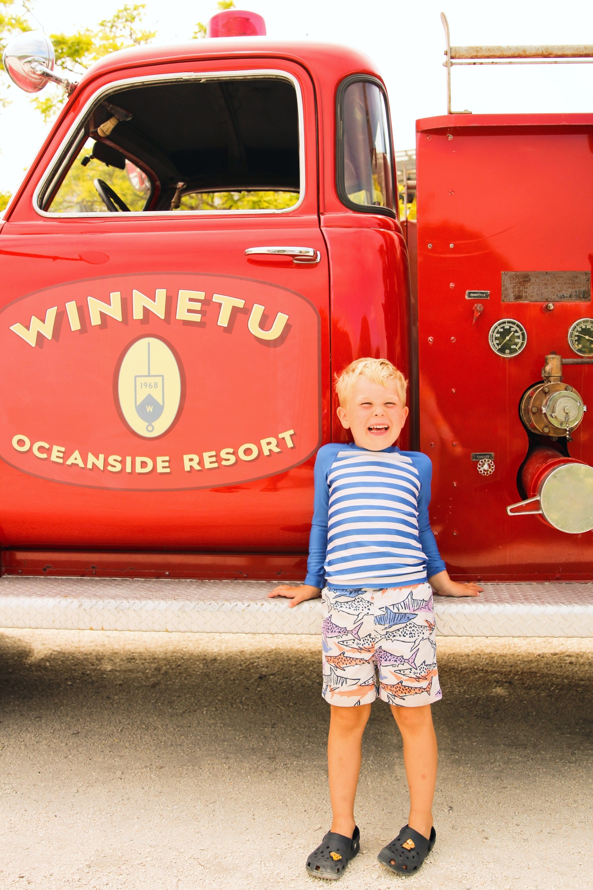 A young child wearing a blue and white striped shirt and patterned shorts stands smiling in front of a vintage red fire truck. The fire truck displays "WINNETU OCEANSIDE RESORT" text on its door along with a circular emblem, and features visible gauges and firefighting equipment on its side panel.