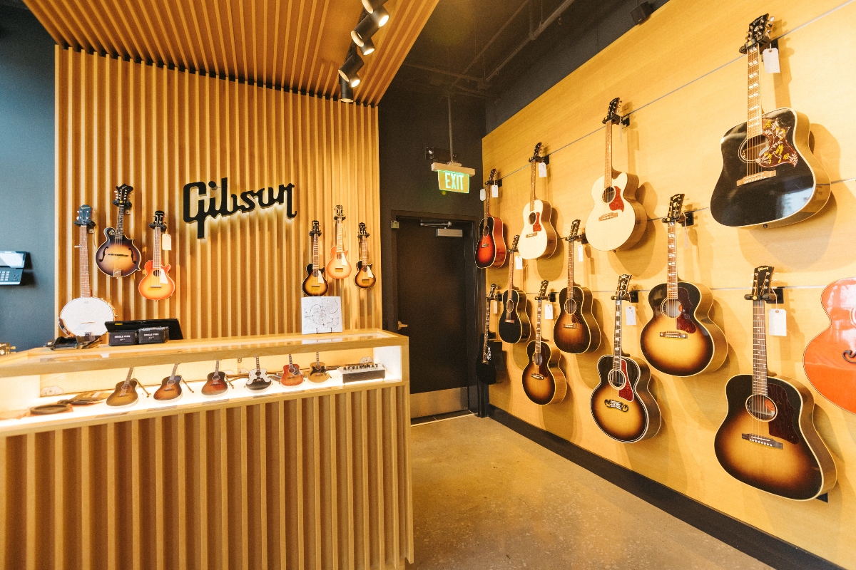 A modern music retail space features a wooden slatted wall and counter displaying the Gibson brand name, with several guitars and ukuleles arranged on the counter beneath wall-mounted instruments. Multiple acoustic guitars in various finishes including sunburst, natural wood, black, and white hang on a pale yellow wall to the right, illuminated by track lighting, with an exit door visible in the background.