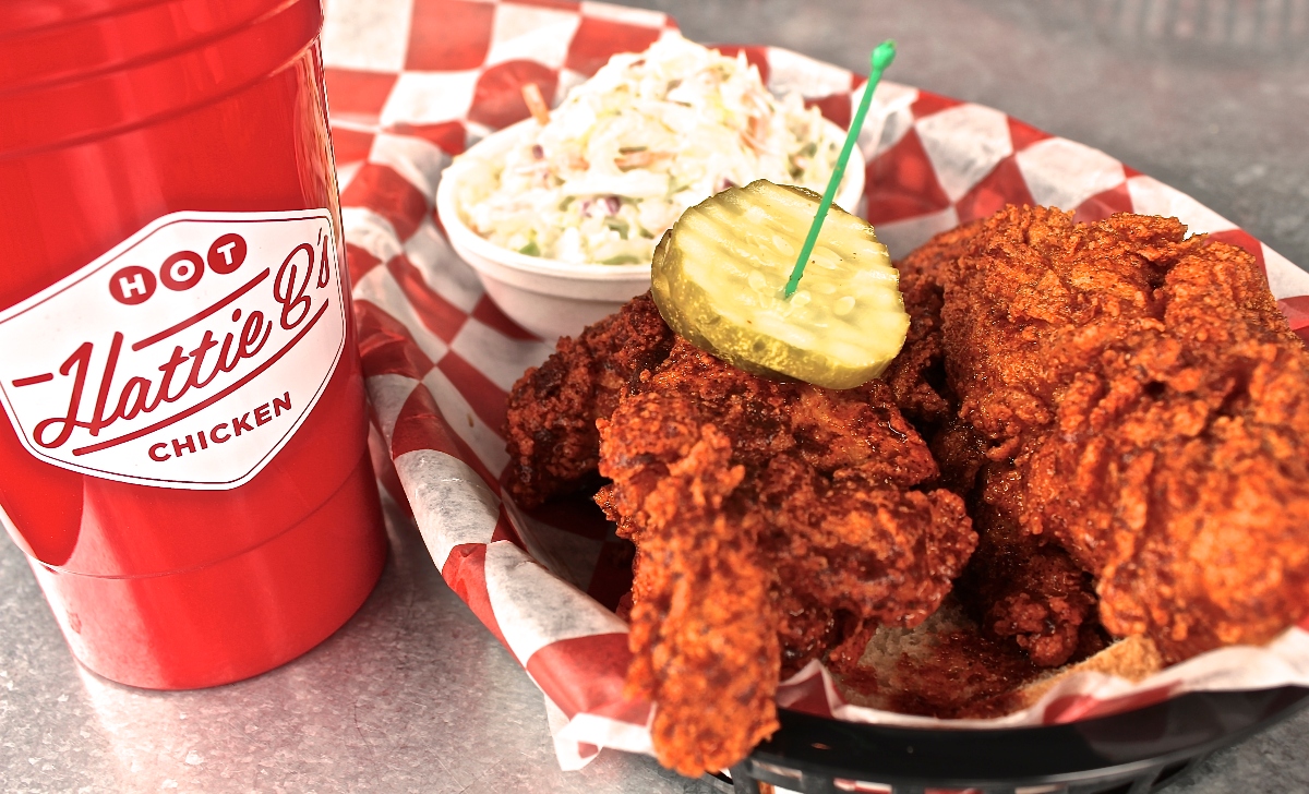 A red branded cup sits beside a basket of crispy fried chicken pieces garnished with a pickle slice secured by a green toothpick, all arranged on red and white checkered paper. A bowl of coleslaw is visible in the background, with the meal displayed on a gray surface.