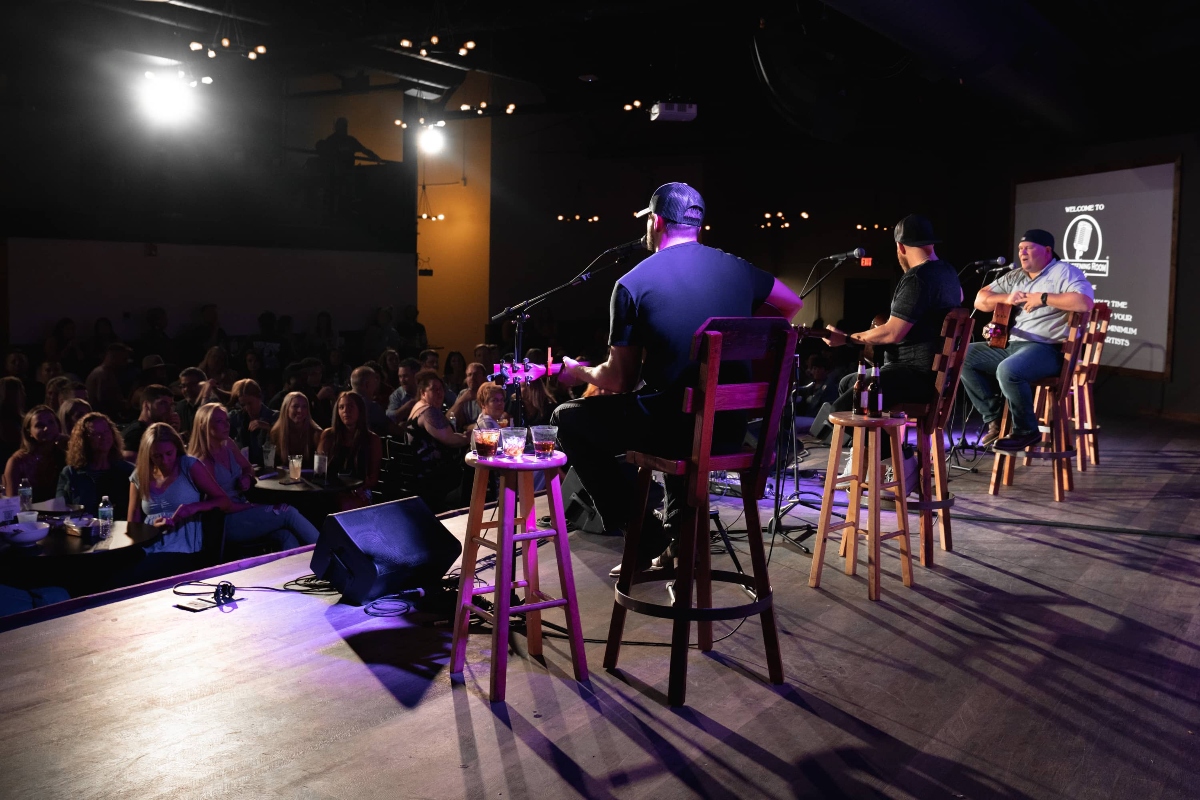 Three musicians seated on bar stools perform an acoustic set on a stage with purple lighting, with microphones and a small table holding drinks positioned in front of them. An audience sits in rows to the left facing the stage, while a projection screen displays text on the right side of the darkened venue.