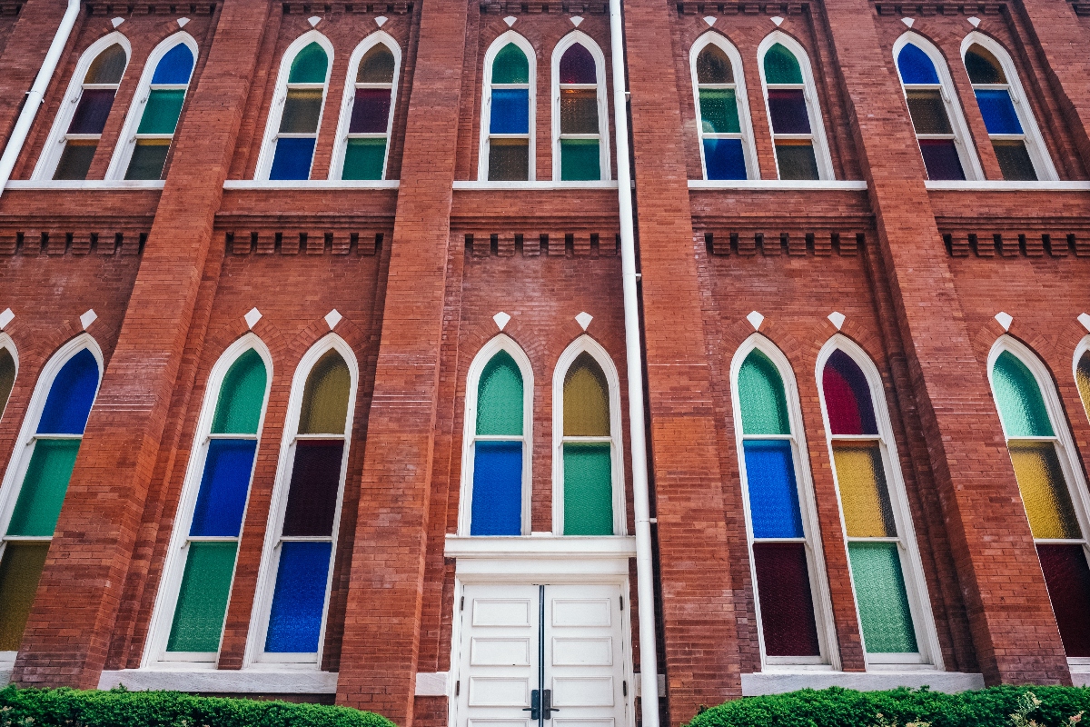 A red brick building facade features two rows of tall Gothic-style arched windows with white frames, each containing multicolored glass panes in shades of blue, green, yellow, orange, and red. A white double door with stone surround sits at ground level, flanked by green shrubbery, with decorative brickwork creating horizontal bands between the window levels.