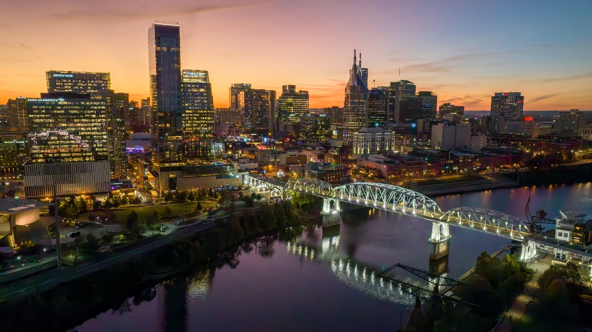 An illuminated white truss bridge spans a river at dusk, with the bridge's lights reflecting on the calm water below. Behind the bridge, a city skyline features numerous high-rise buildings lit against a gradient sky transitioning from orange and pink near the horizon to deep blue above.