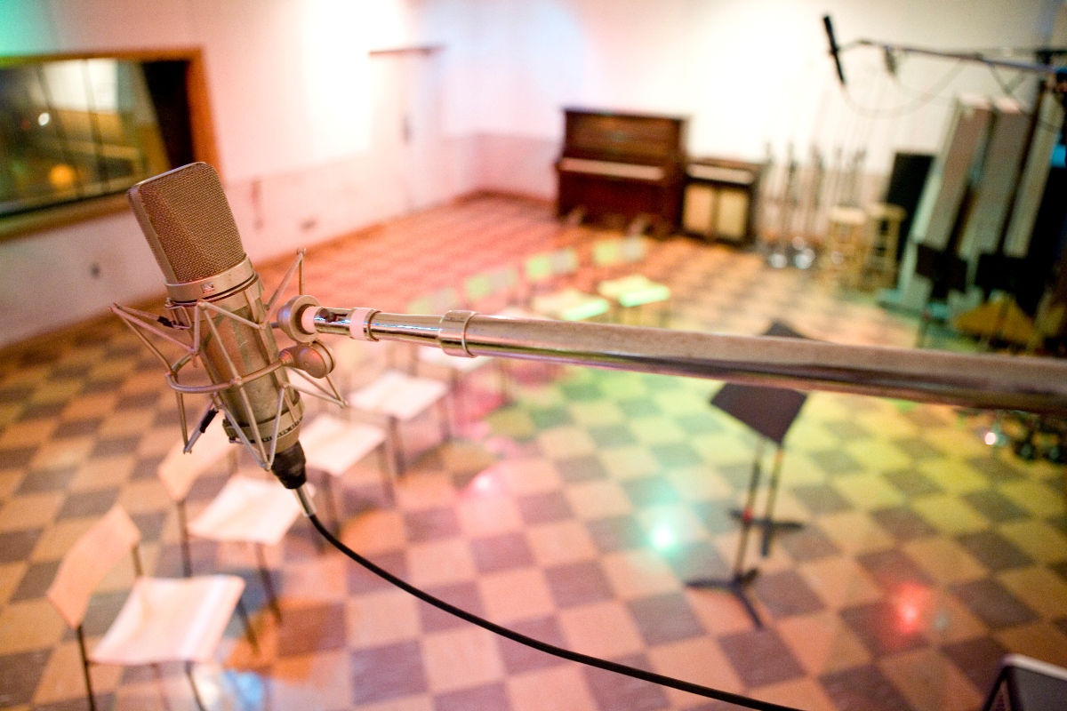 A professional condenser microphone mounted on a shock mount sits in the foreground, positioned on a boom arm with its cable visible. The blurred background shows a recording studio space with a piano, control desk with papers and equipment, and a colorful patterned floor.