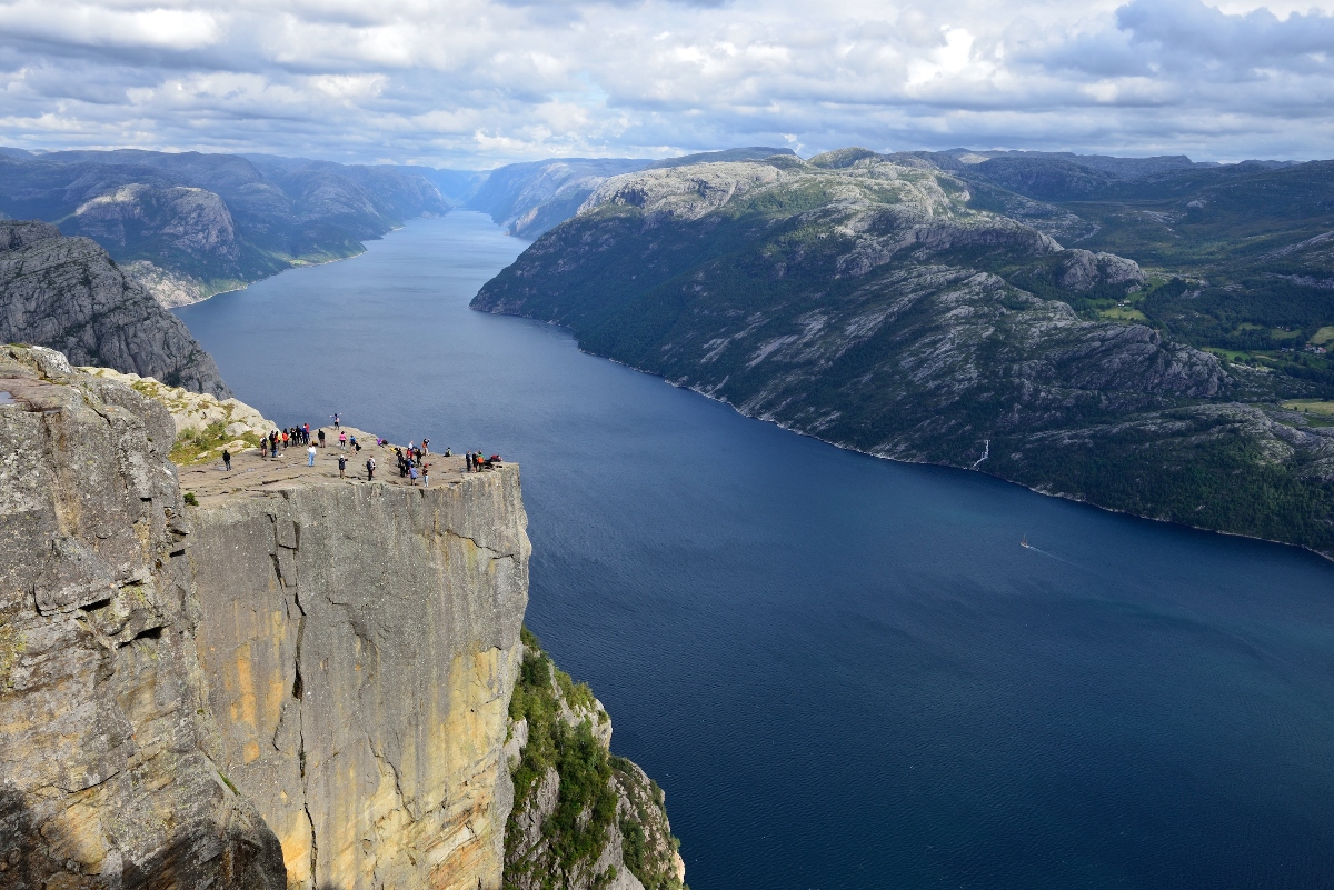 A large group of people stand on a flat-topped cliff edge that juts out dramatically over a deep blue fjord hundreds of meters below. The vertical cliff face shows gray rock with yellow and orange mineral staining, while steep mountains with exposed rock and patches of green vegetation rise on all sides of the narrow fjord. A cloudy sky stretches across the distant mountain peaks that frame the water corridor.