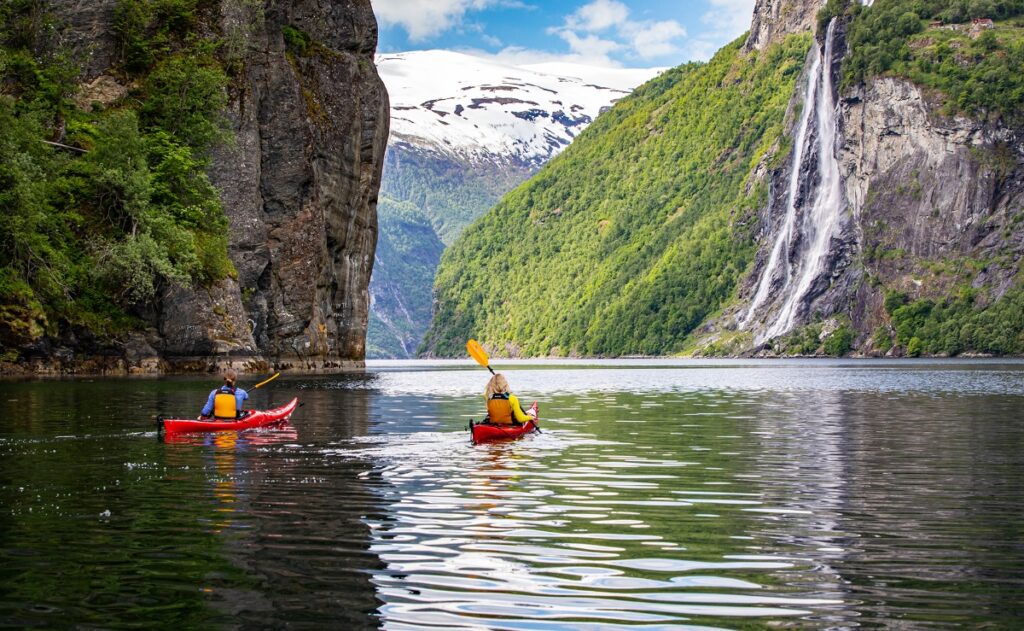 Two kayakers in red kayaks wearing orange life jackets paddle on calm water surrounded by steep mountain cliffs densely covered with green vegetation. A tall waterfall cascades down the gray rock face on the right side of the fjord, while a massive rock overhang protrudes from the left cliff. A snow-covered mountain peak is visible in the distance beneath a partly cloudy blue sky.