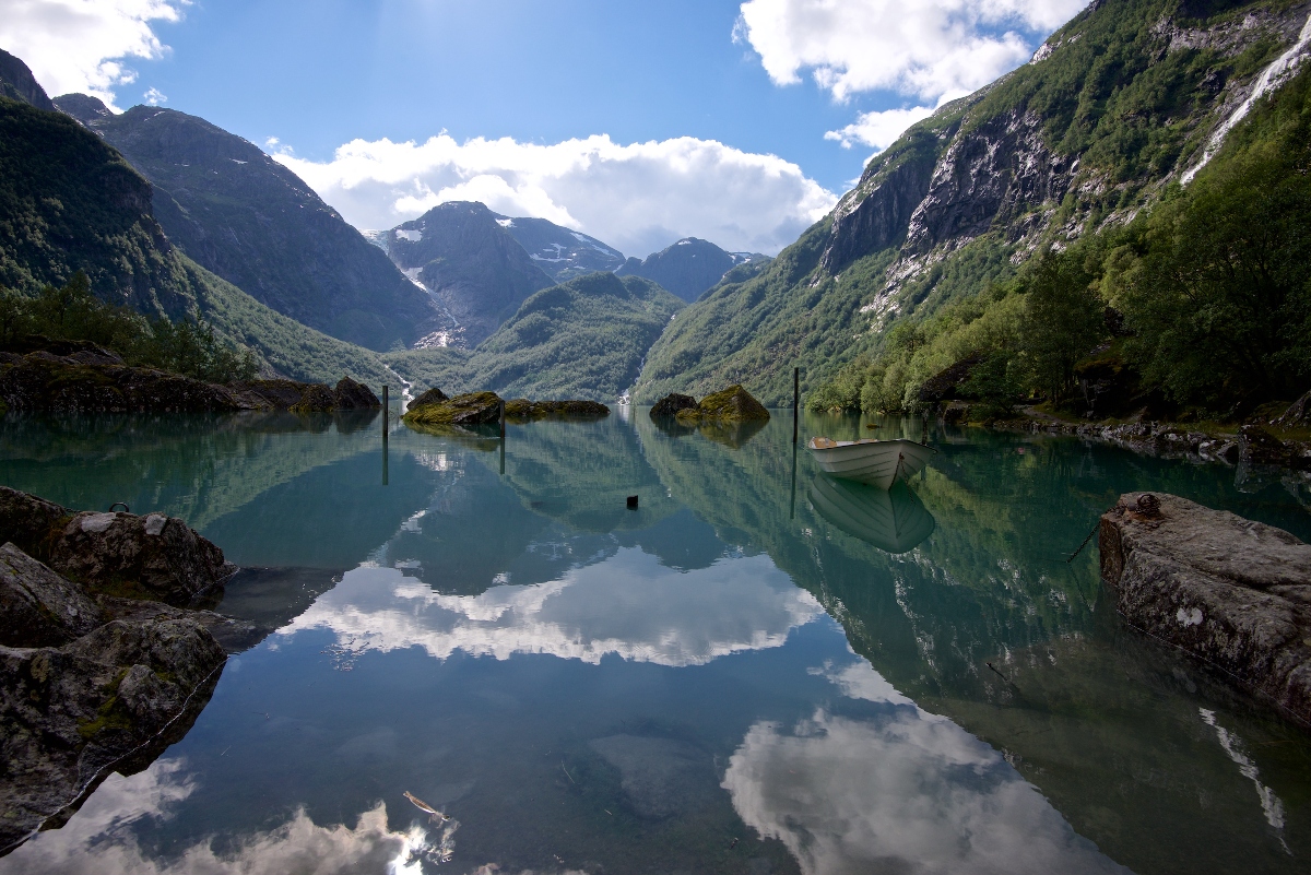 A tranquil fjord in Norway reflects the surrounding green mountains, blue sky, and scattered clouds like a mirror. A small white rowboat floats quietly near the shore, framed by rocky edges and lush forested slopes.