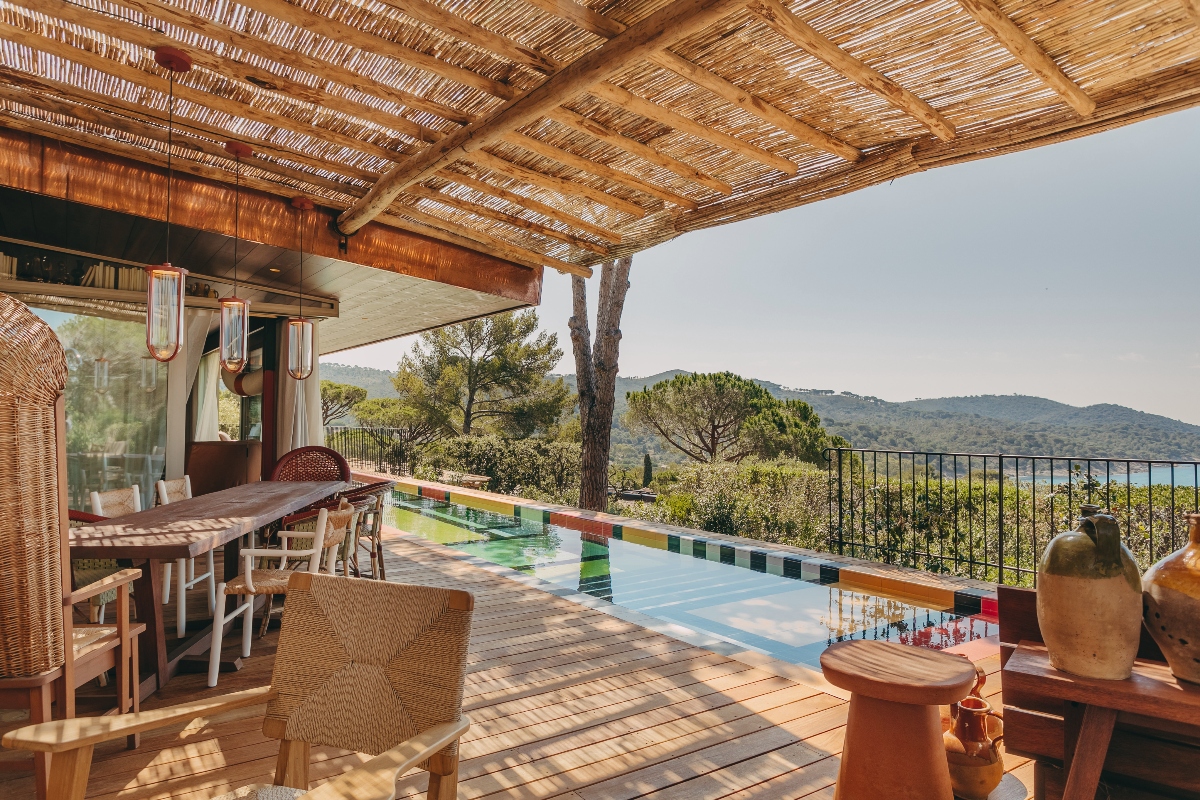 A wooden deck terrace features wicker furniture arranged under a bamboo-roofed pergola, with a colorful tiled swimming pool running along one edge. The outdoor space overlooks a mountainous landscape with trees and vegetation, enclosed by a black metal railing.