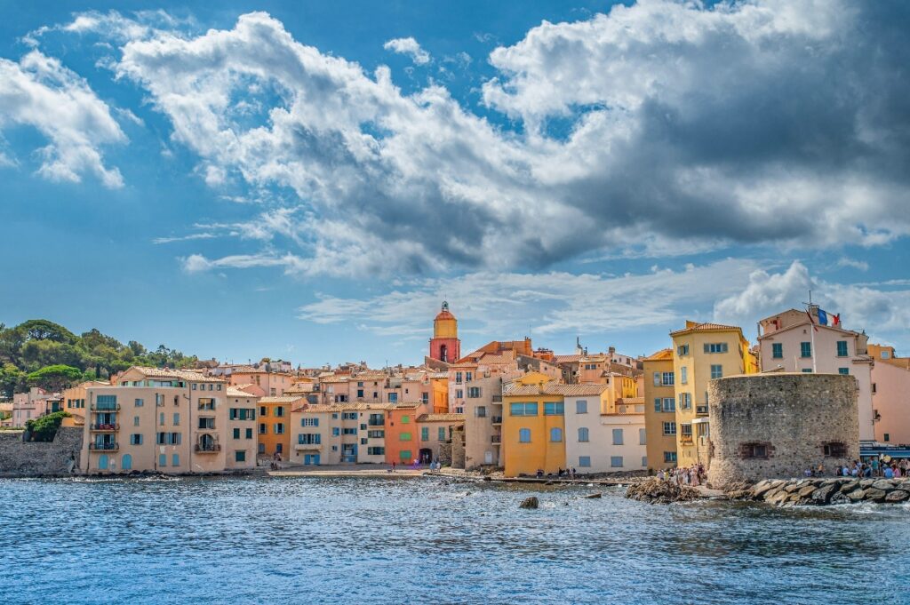 A Mediterranean coastal town displays rows of colorful multi-story buildings in shades of yellow, orange, pink, and cream along the waterfront, with a prominent bell tower rising above the rooflines. The scene is viewed across calm blue water under a dramatic sky filled with white and gray clouds, with a stone fortification visible on the right side and wooded hills in the background.