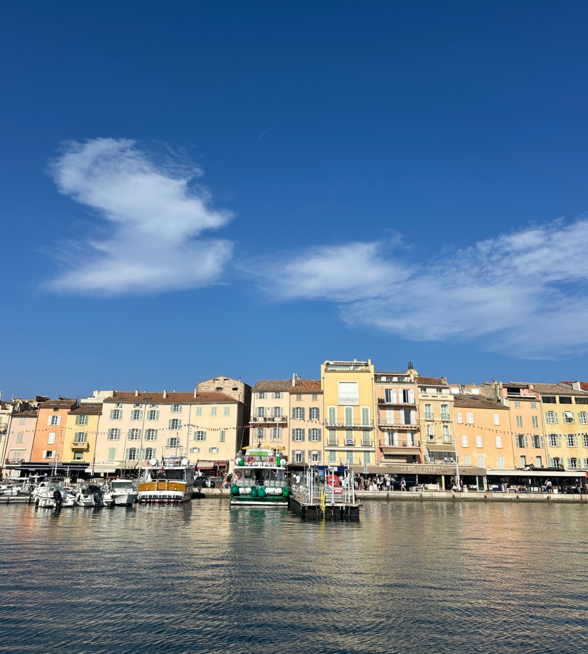 A row of colorful multi-story buildings in warm tones of yellow, peach, and cream line a waterfront harbor, with shops and restaurants visible at street level and people walking along the promenade. Several small boats and pleasure craft are moored in the calm blue water under a bright blue sky dotted with white wispy clouds.