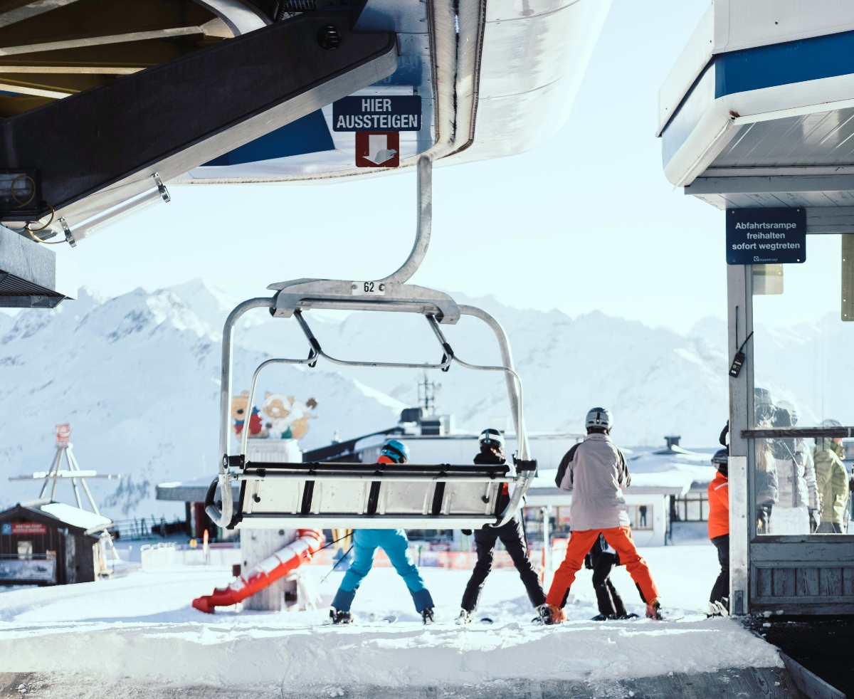 A ski lift station with a sign reading "HIER AUSSTEIGEN" (exit here) is shown with a chairlift passing overhead carrying passengers. Several skiers, including children wearing brightly colored ski pants in orange, blue, and black, are disembarking or walking near the lift station. Snow-covered mountain peaks are visible in the misty background behind the ski resort infrastructure.